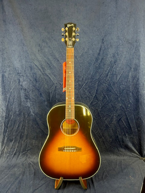 Sunburst finish acoustic guitar with a rounded body, six strings, and a dark-to-light gradient on the front, standing upright on a wooden stand.
