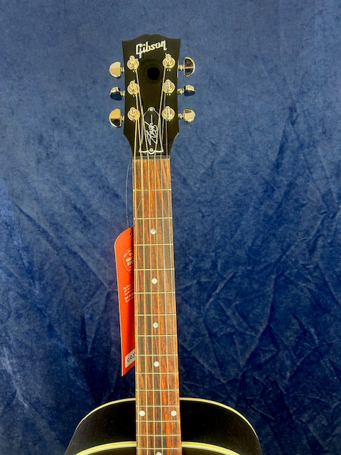 Gibson acoustic guitar with a dark headstock, gold tuning pegs, and a rosewood fretboard against a textured blue background. Red tag hangs near tuners.