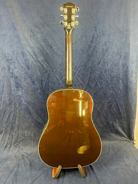 A glossy brown acoustic guitar is displayed upright on a stand, showing its back and neck against a dark blue fabric background.