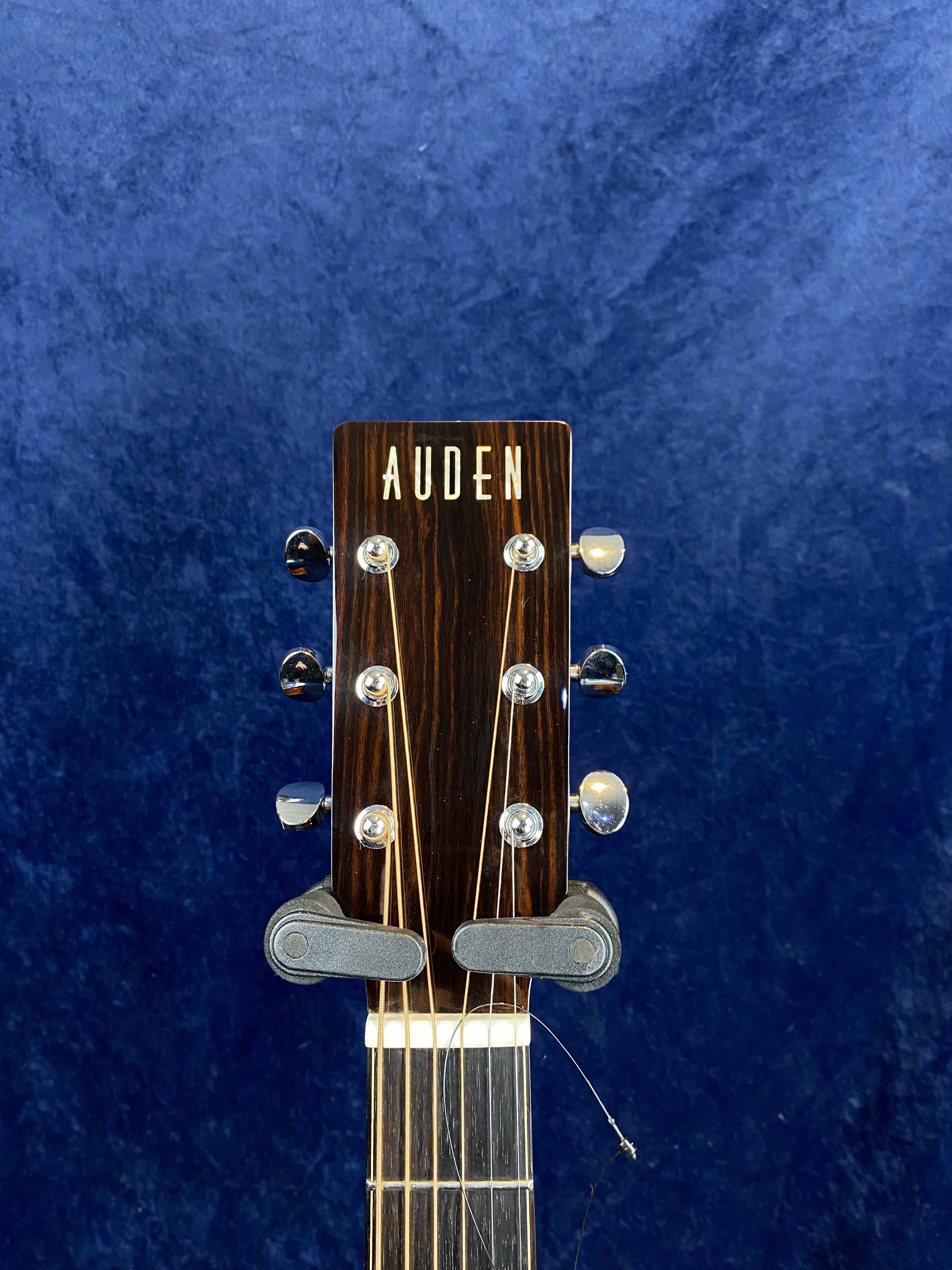 The headstock of an Auden acoustic guitar with six tuning pegs and strings, set against a dark blue background.