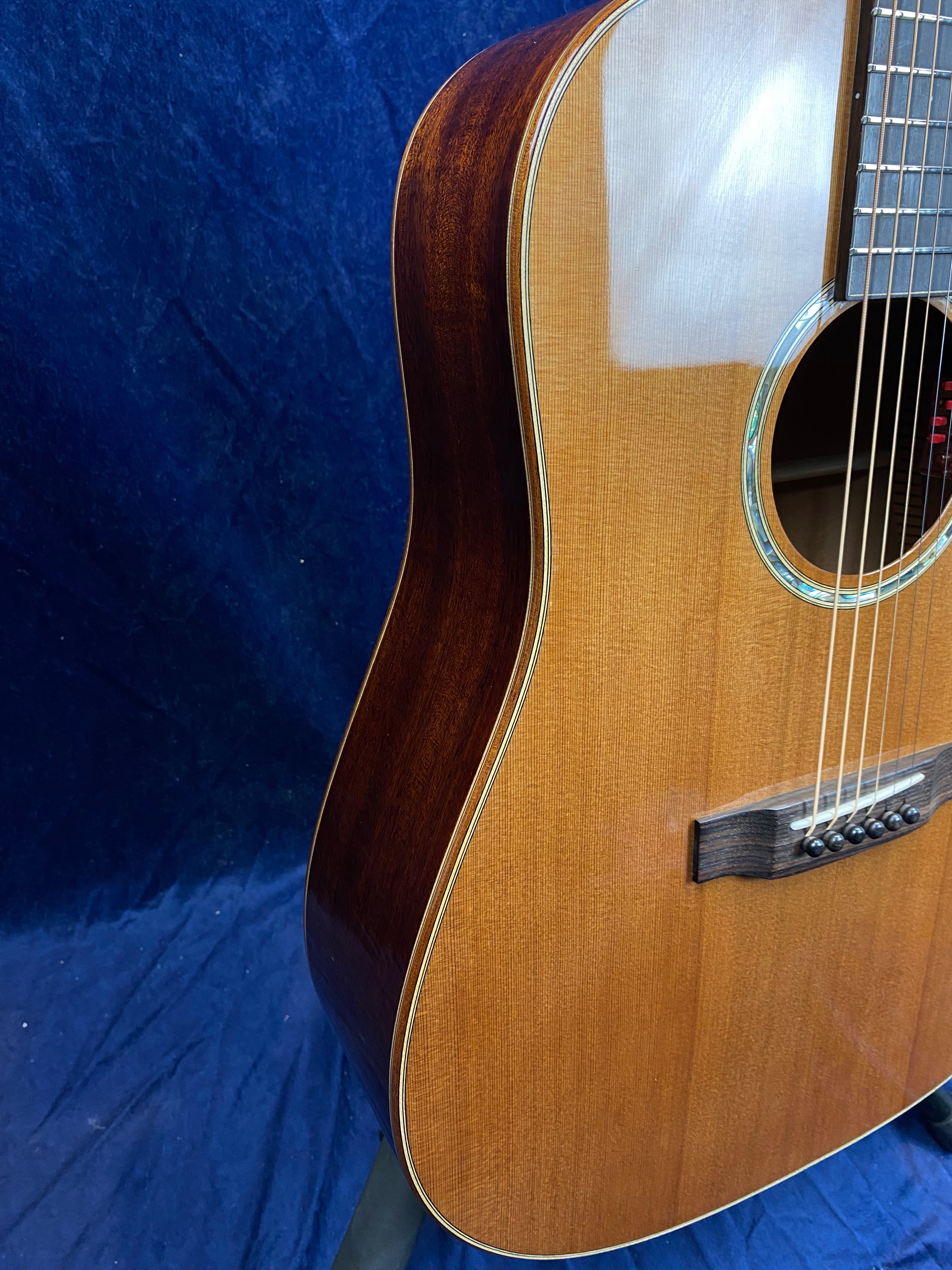 Acoustic guitar with a glossy light brown top, dark brown sides, and an abalone-trimmed soundhole against a blue background.