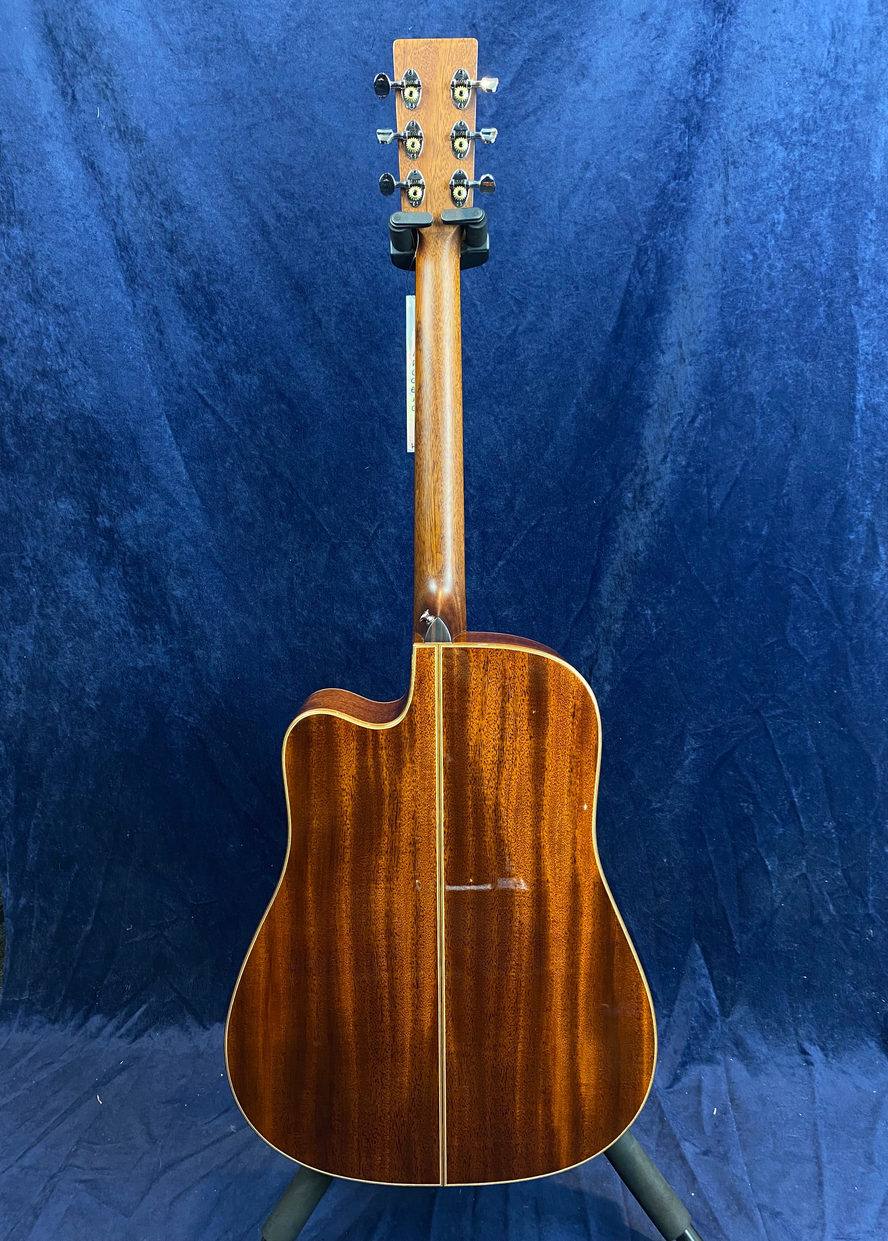 Back view of an acoustic guitar with rich brown wood, gold outline, and six tuning pegs, standing upright against a dark blue background.