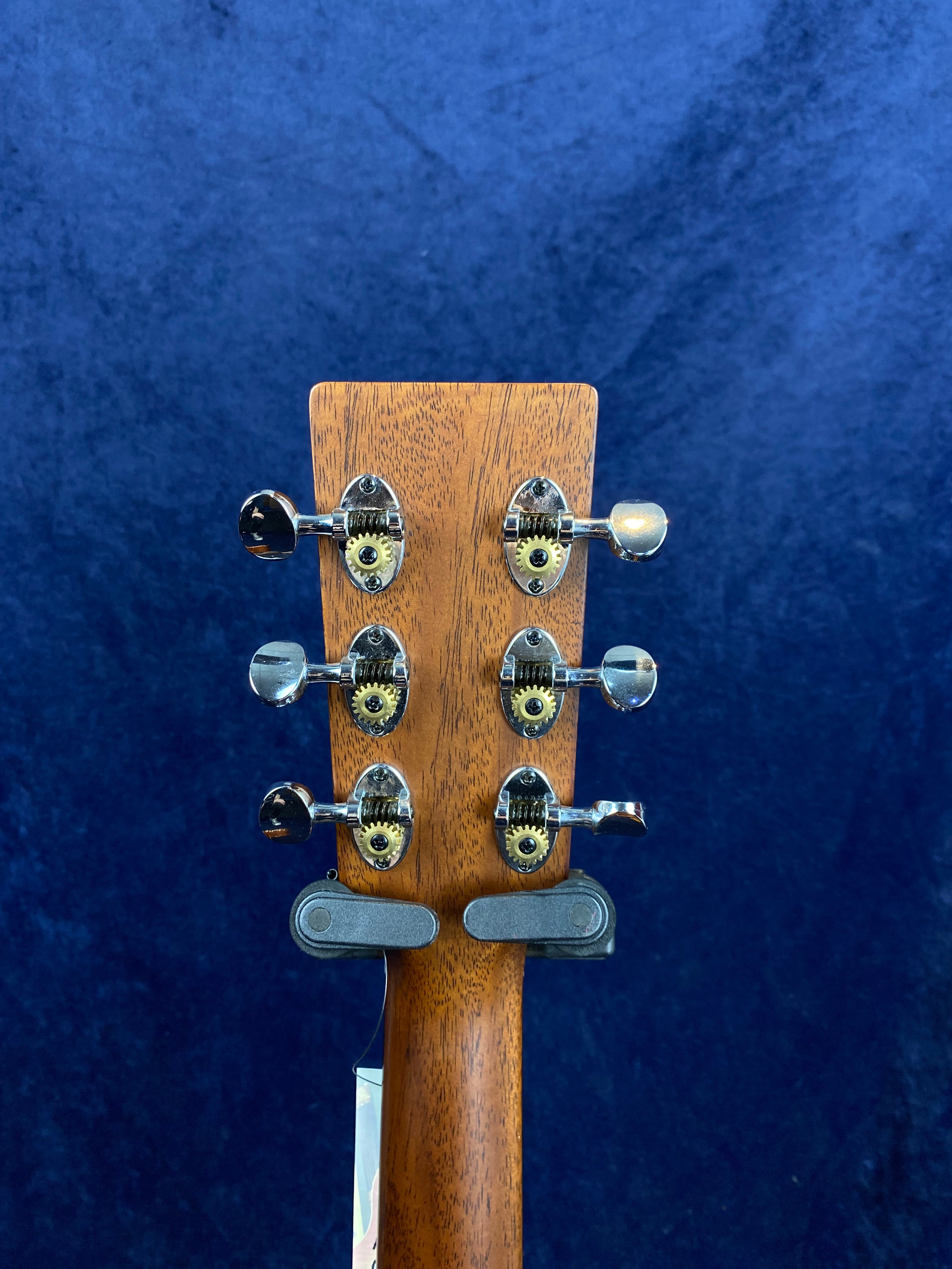 Six metal tuning pegs with exposed gears on the headstock of a wooden acoustic guitar, set against a dark blue background.