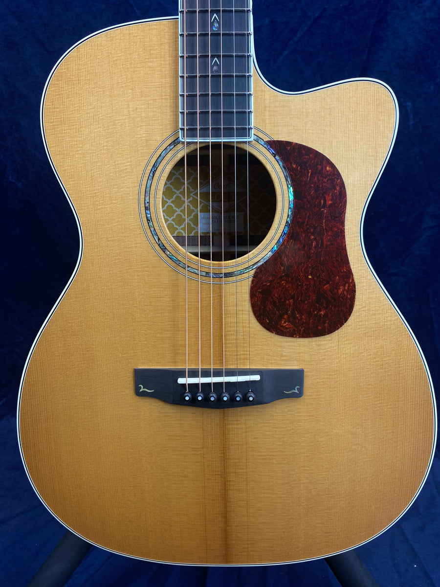 Light brown acoustic guitar with a cutaway body, dark tortoiseshell pickguard, abalone rosette, and six strings with pearl inlays on the fretboard.