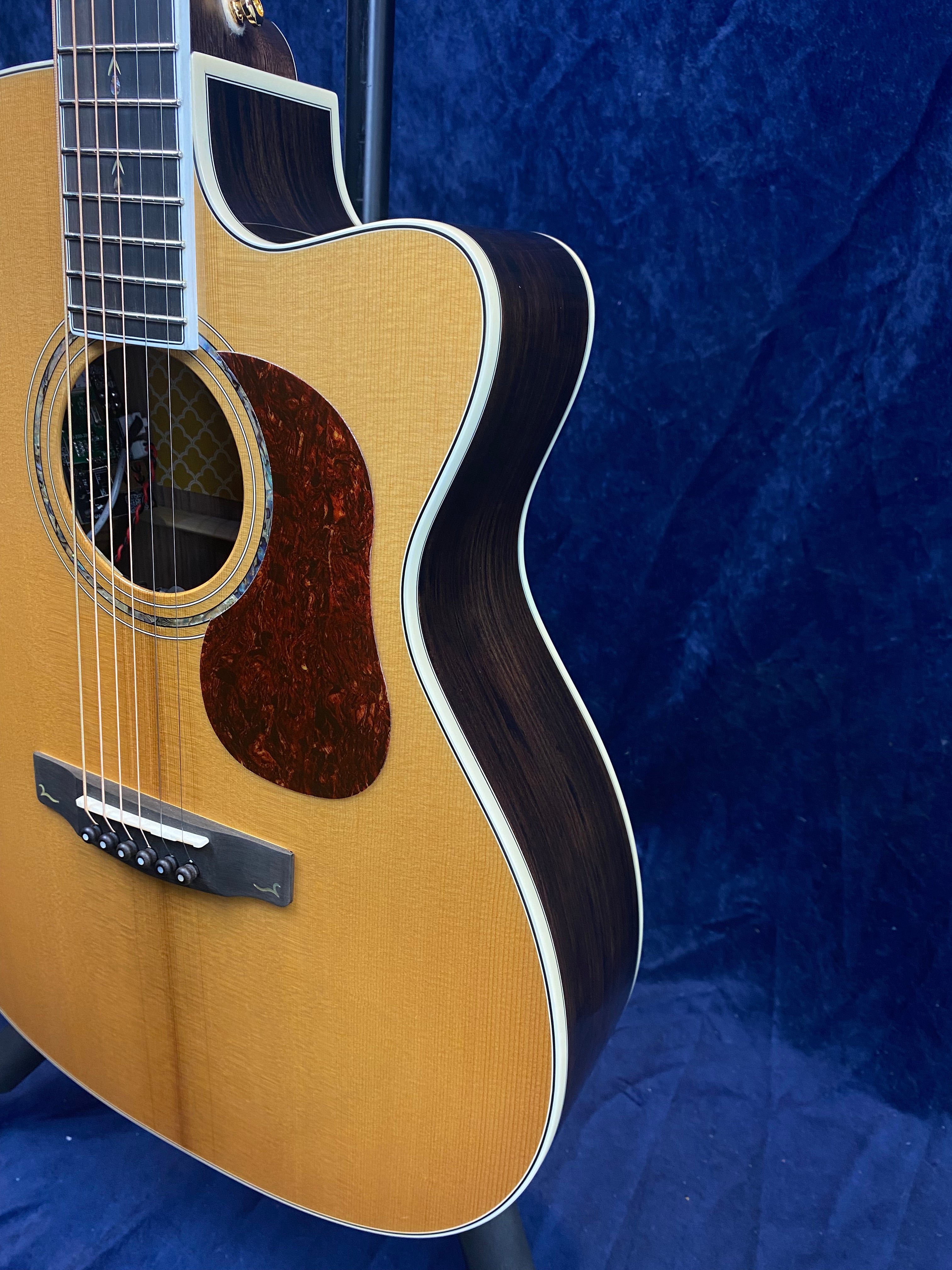 Cutaway acoustic guitar with a light wood top, dark wood sides, white binding, and a reddish-brown pickguard against a dark blue background.