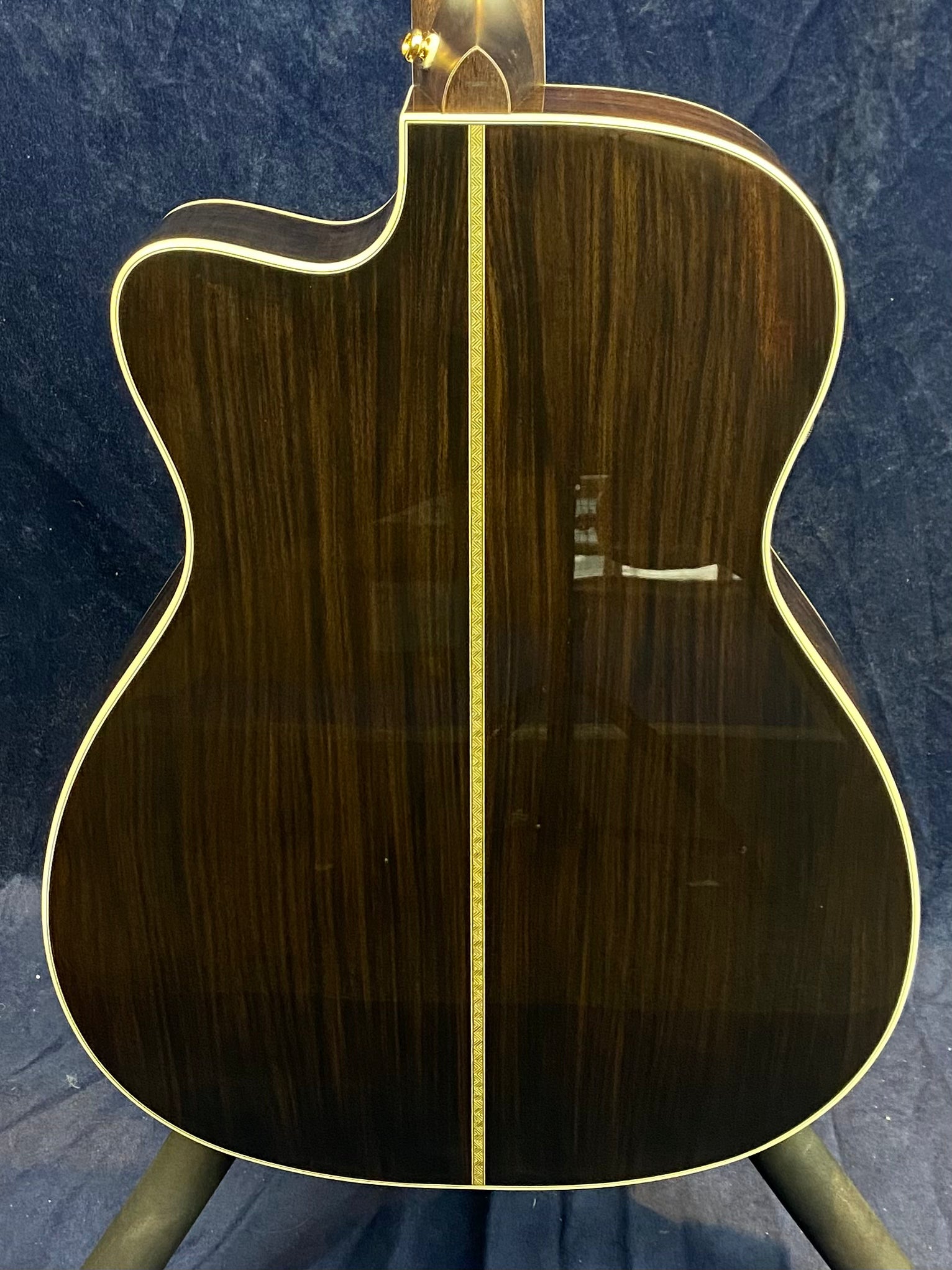 Back of a dark wood acoustic guitar with a decorative central strip and light binding outlining the body against a dark background.