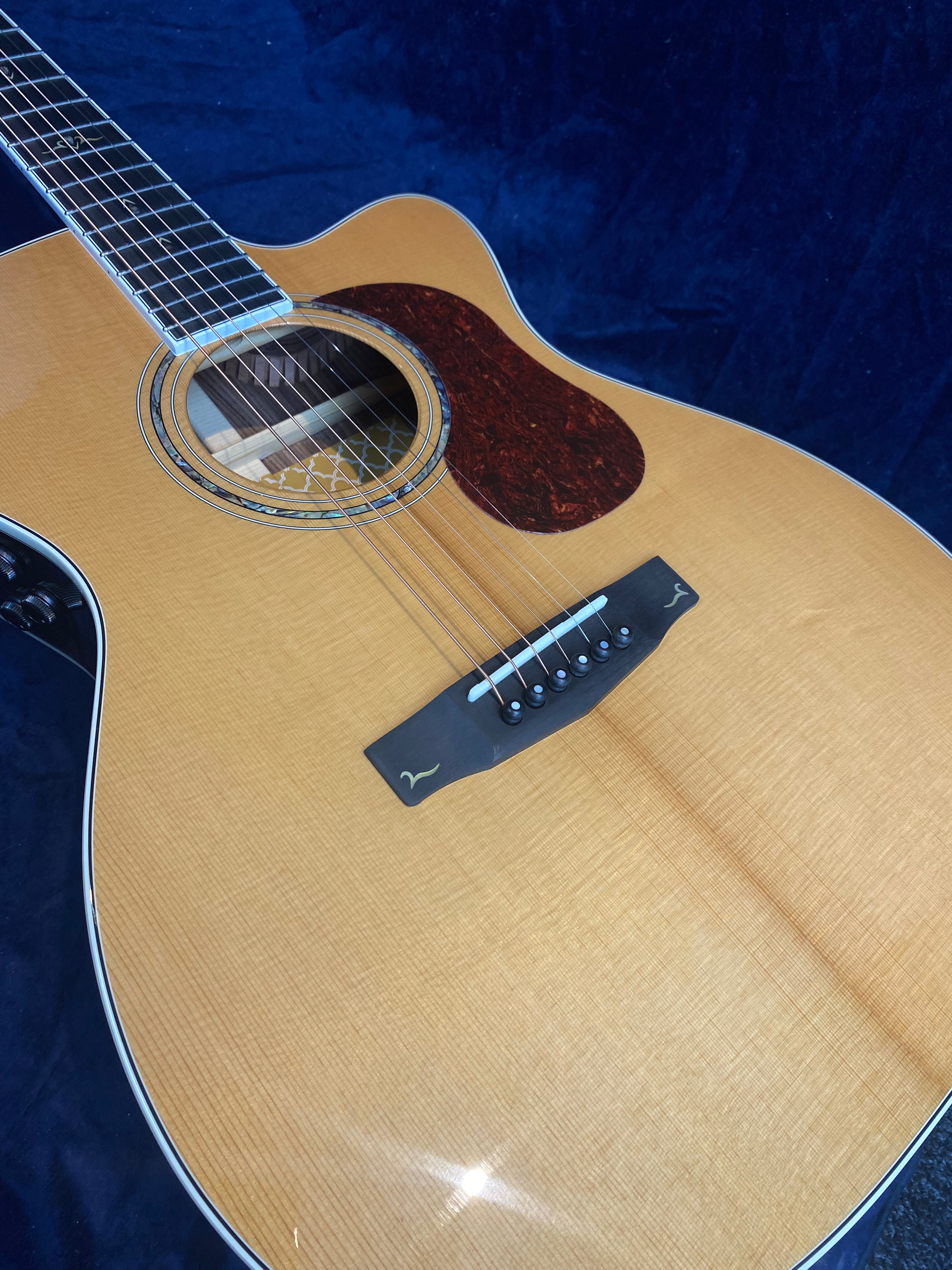Natural wood finish acoustic guitar with a tortoiseshell-style pickguard, black bridge, and white outline accents against a dark blue background.