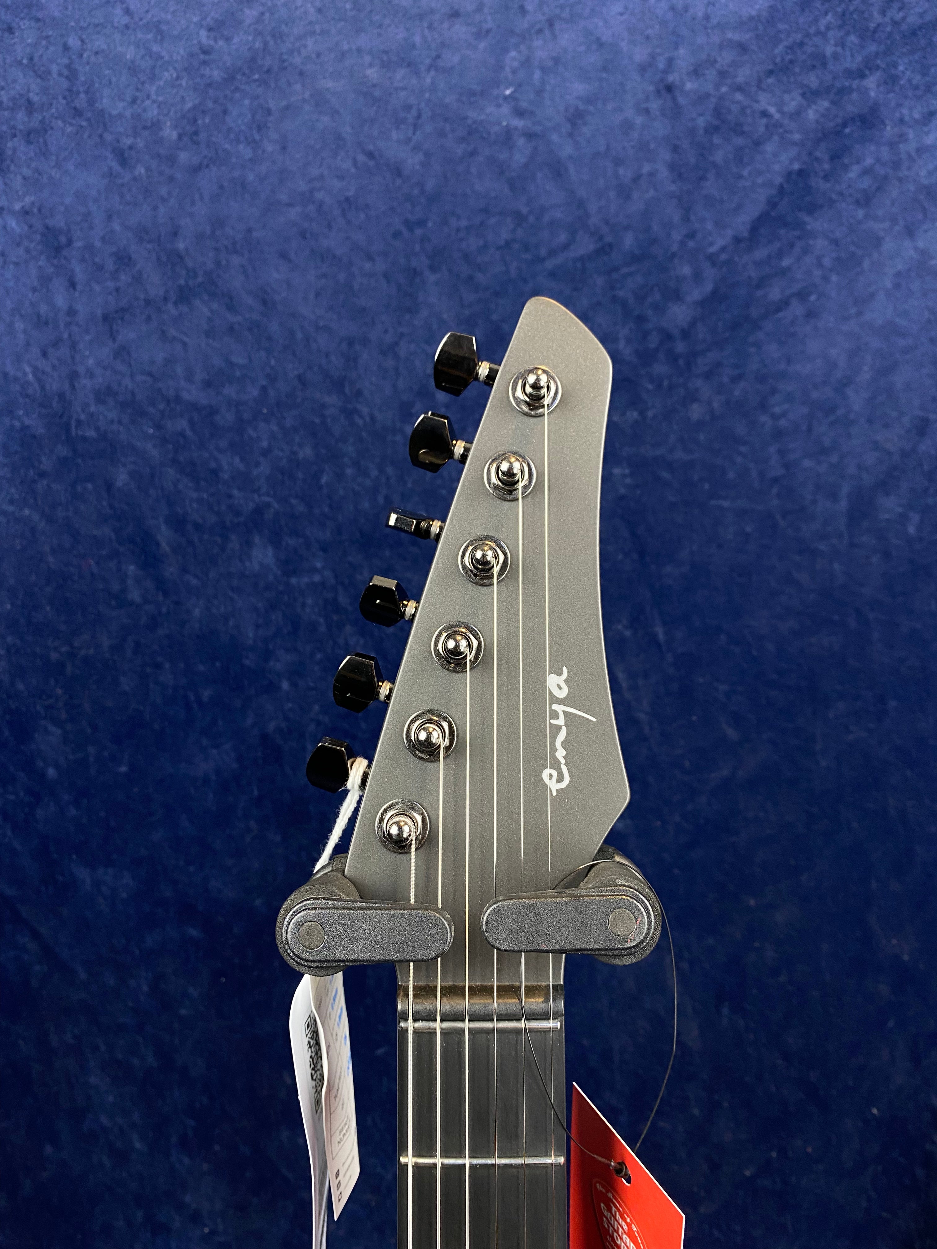 Metallic gray electric guitar headstock with six black tuning pegs, white "enya" logo, and attached hang tags against a dark blue background.