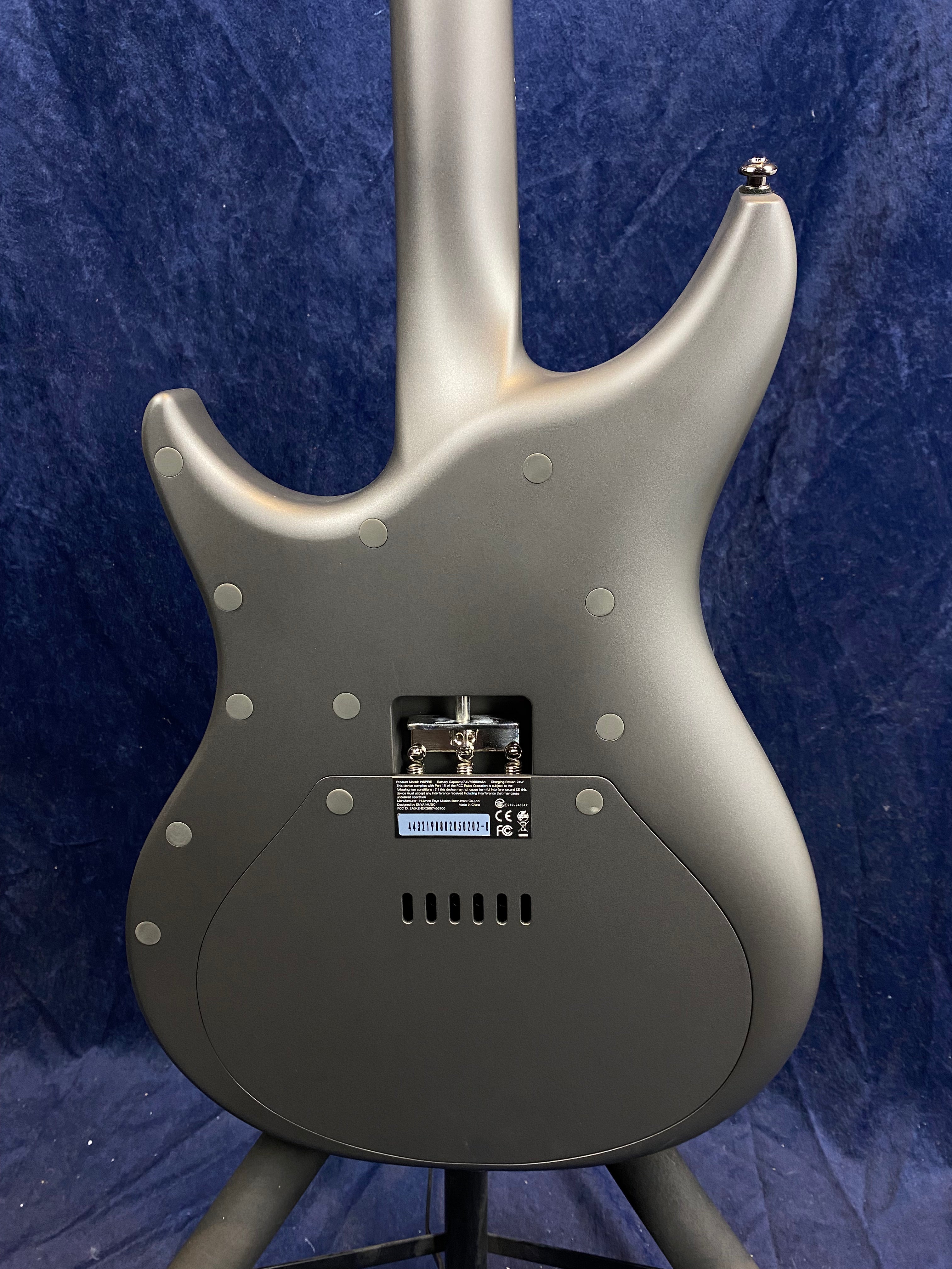 Matte gray electric guitar back showing a contoured body, bolt-on neck, large electronics cover, tremolo cavity, and visible screws on a stand.