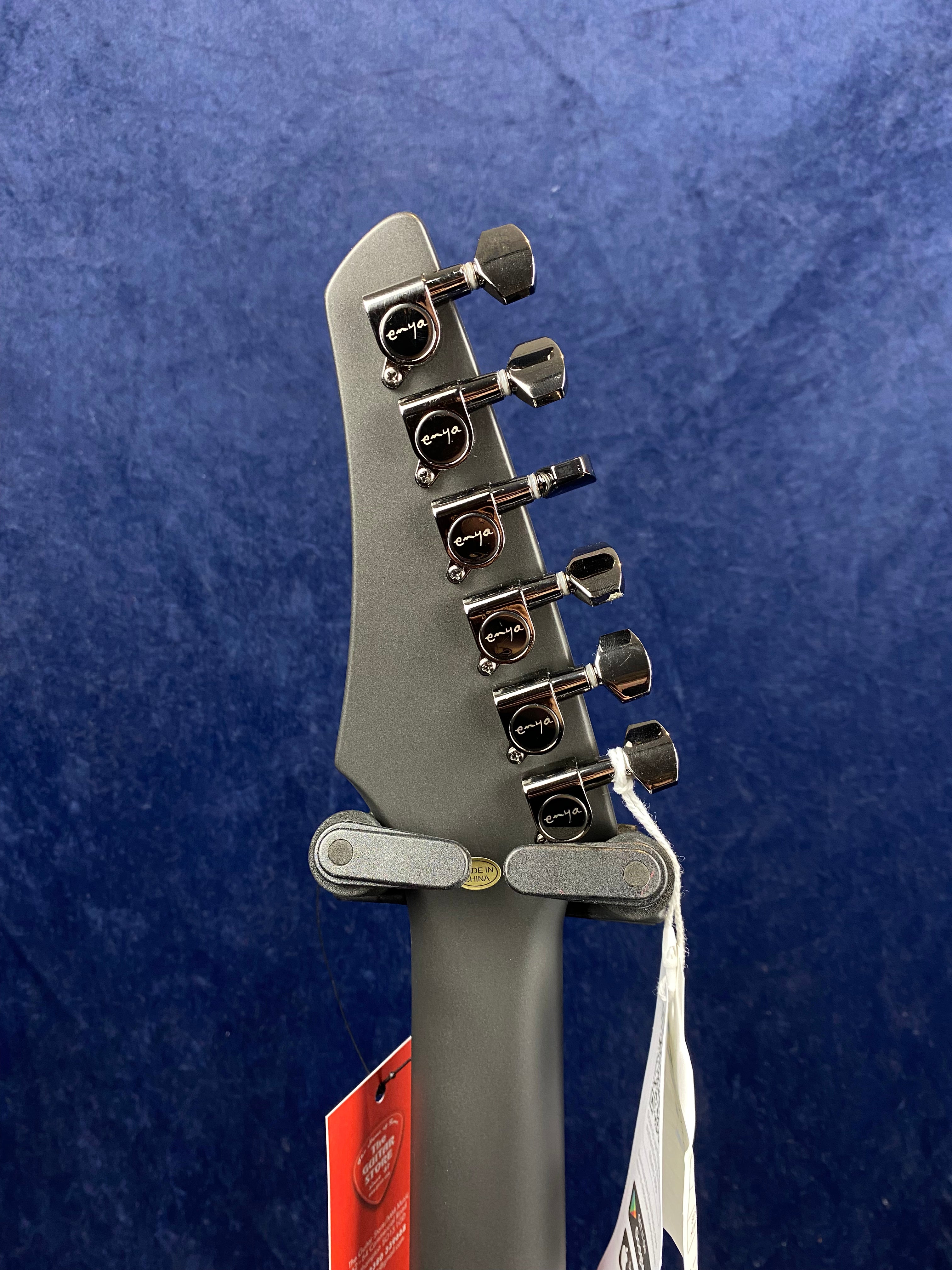 Six tuning pegs with "enya" logos are mounted on a matte gray electric guitar headstock, seen from the back, with tags hanging from the neck.