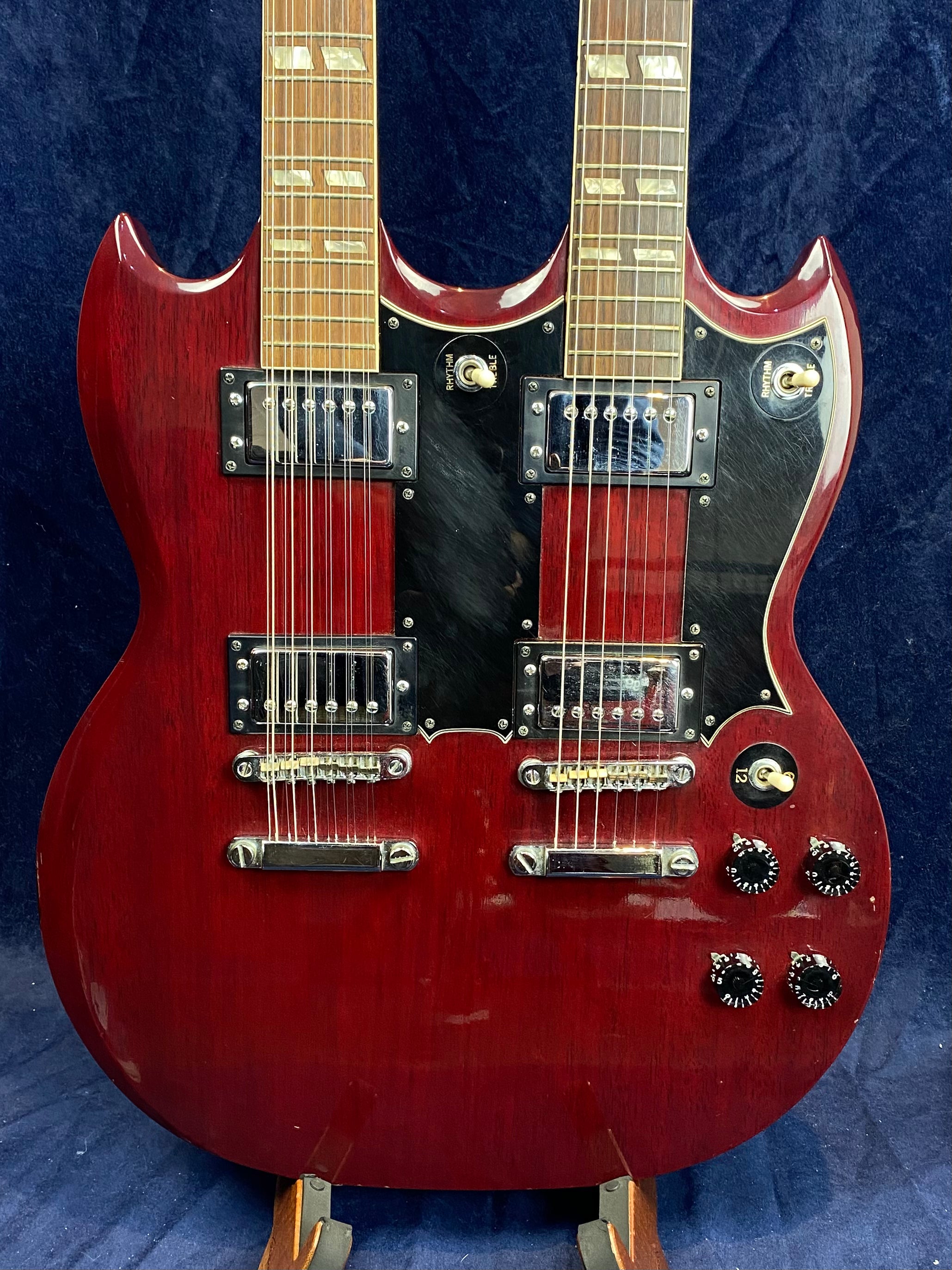 Cherry red double-neck electric guitar with two sets of pickups, knobs, and switches, mounted on a stand against a dark background.