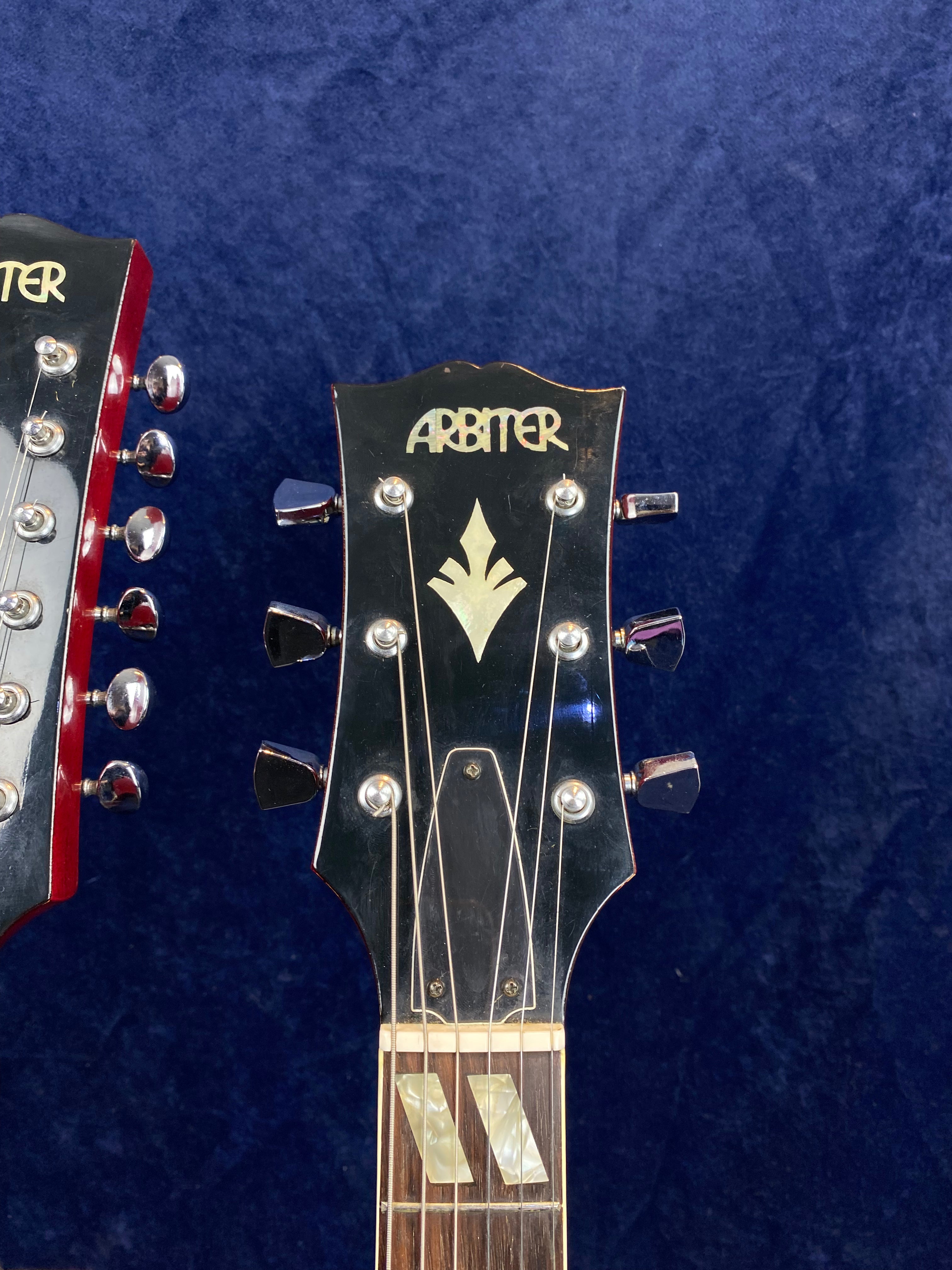 Two electric guitar headstocks with "Arbiter" inlaid logo and decorative inlay, metal tuning pegs, and a dark glossy finish against a blue background.