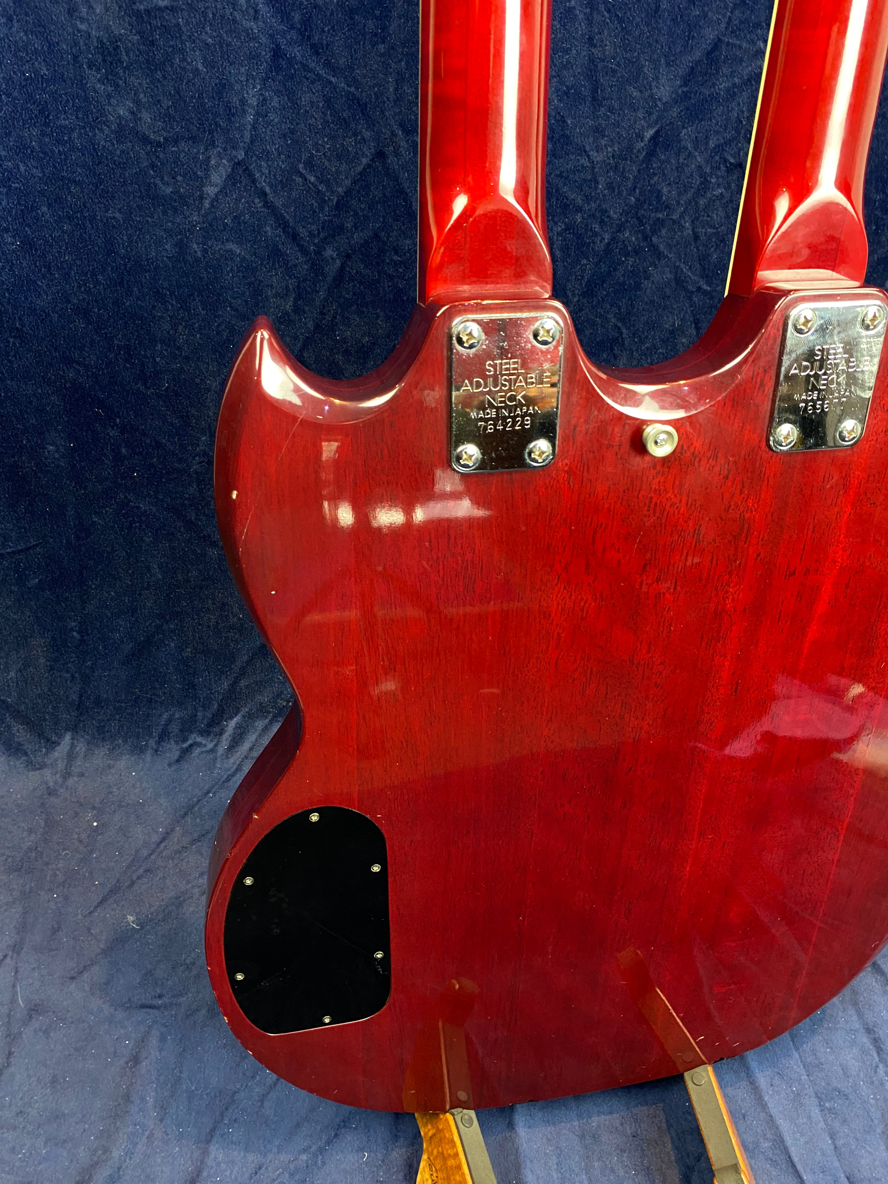 Red double-neck electric guitar, back view, showing two neck plates labeled "Steel Adjustable Neck Made in Japan" and a black electronics cover.