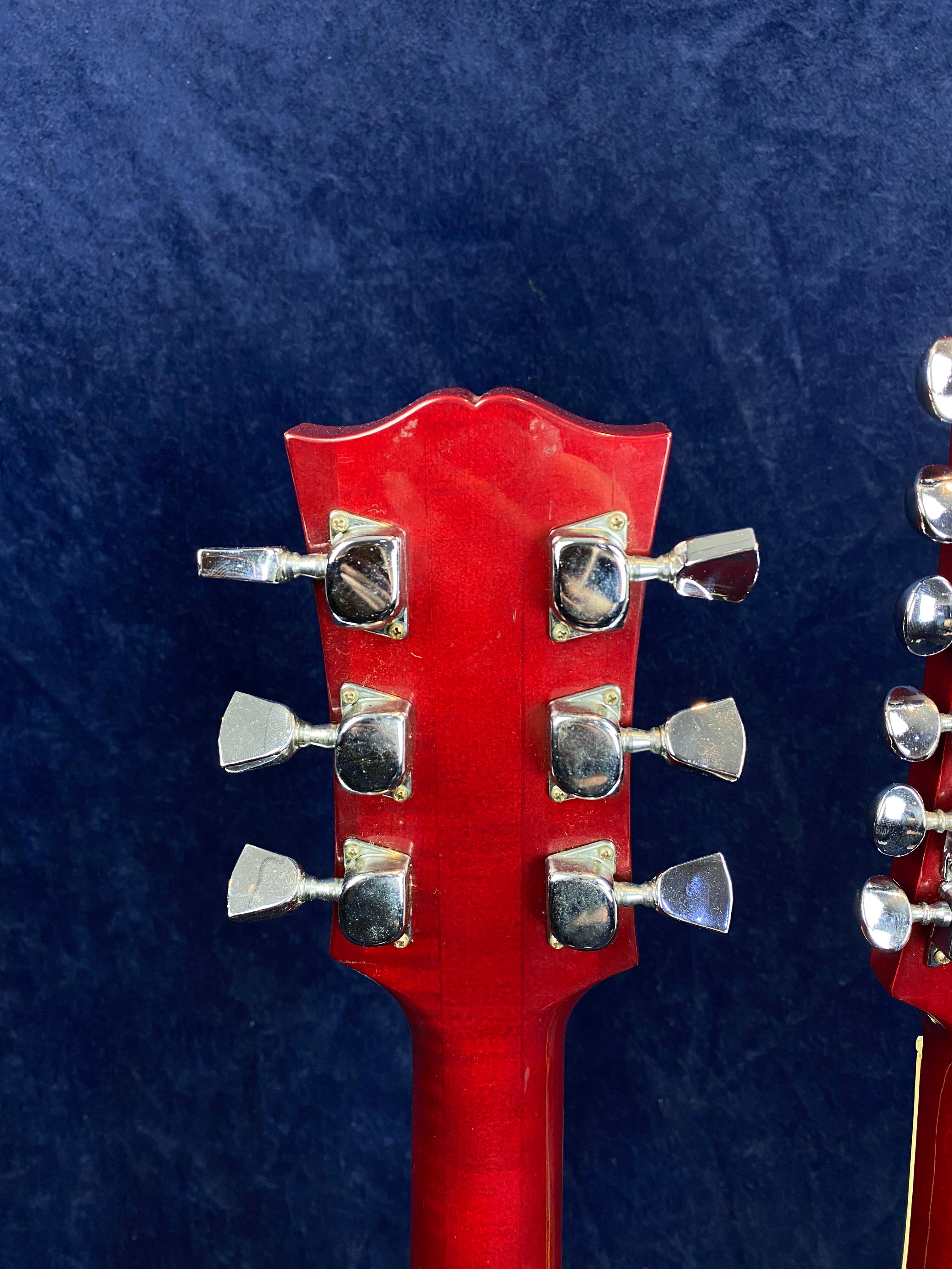 Back of a red electric guitar headstock with six silver tuning pegs, flared top edge, against a dark blue background.