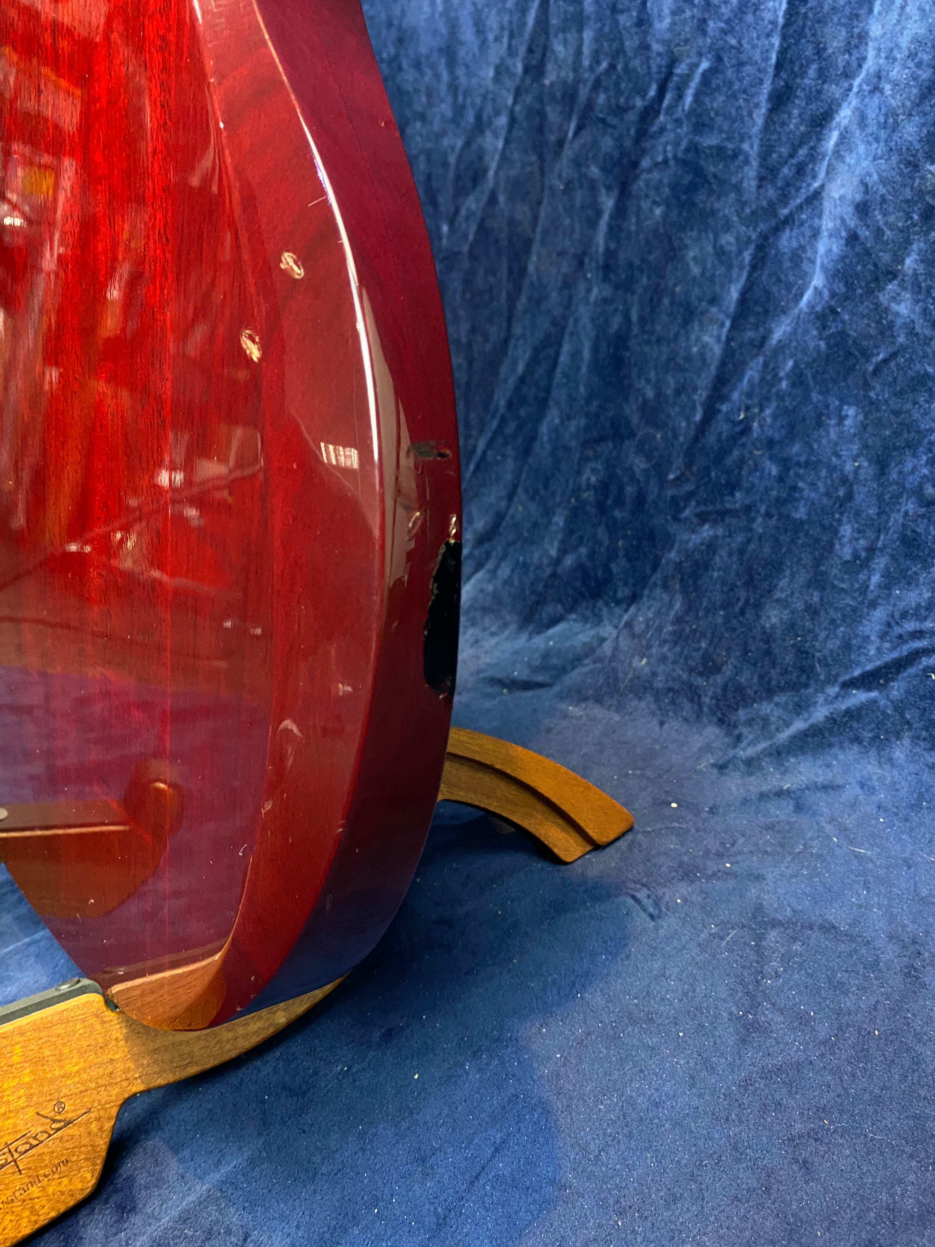 Red electric guitar body with visible scratches and dings, resting on a wooden stand against a blue fabric background.