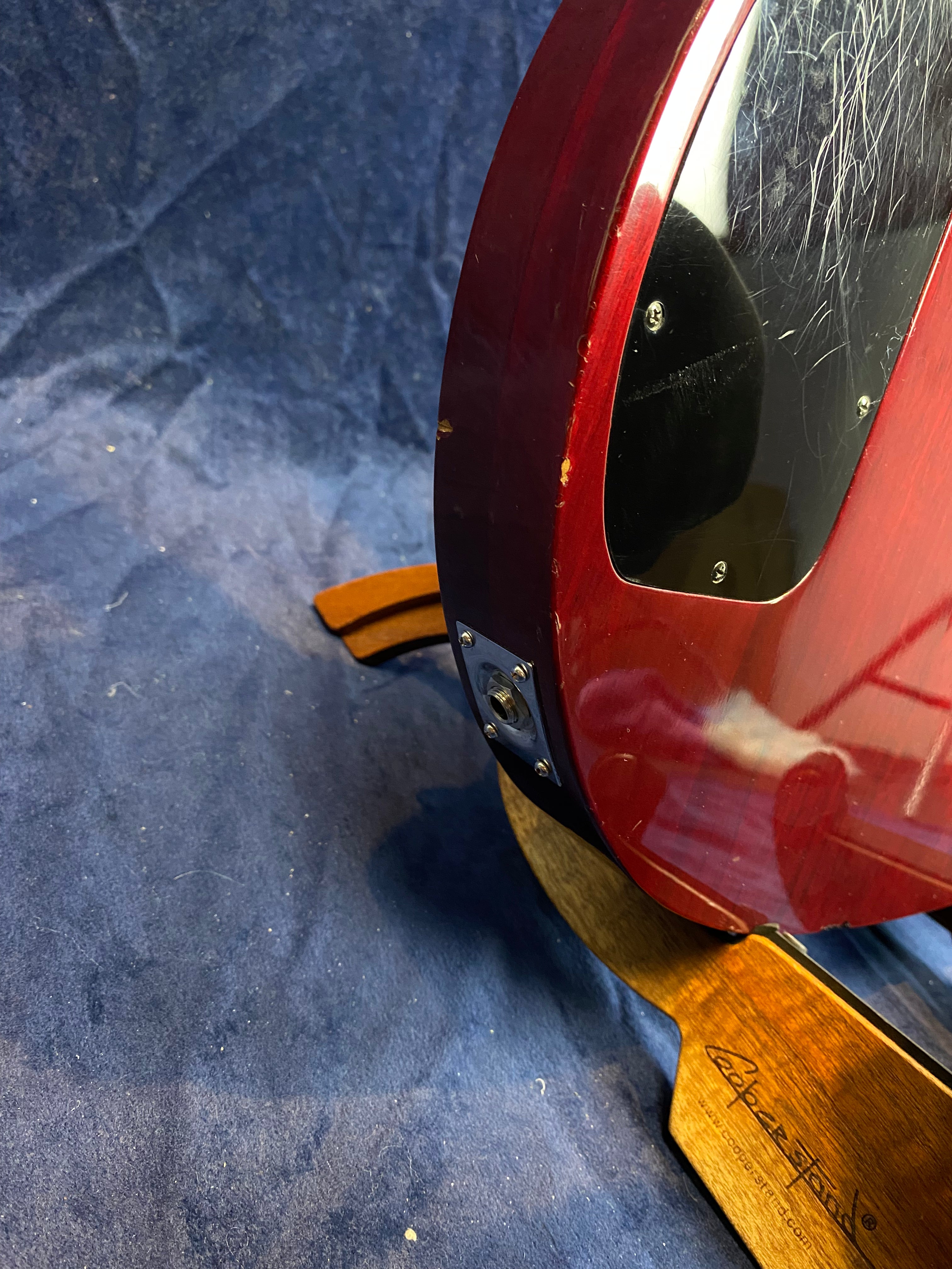 Back edge of a red electric guitar with scratches and small dings, shown on a wooden stand against a blue fabric background.