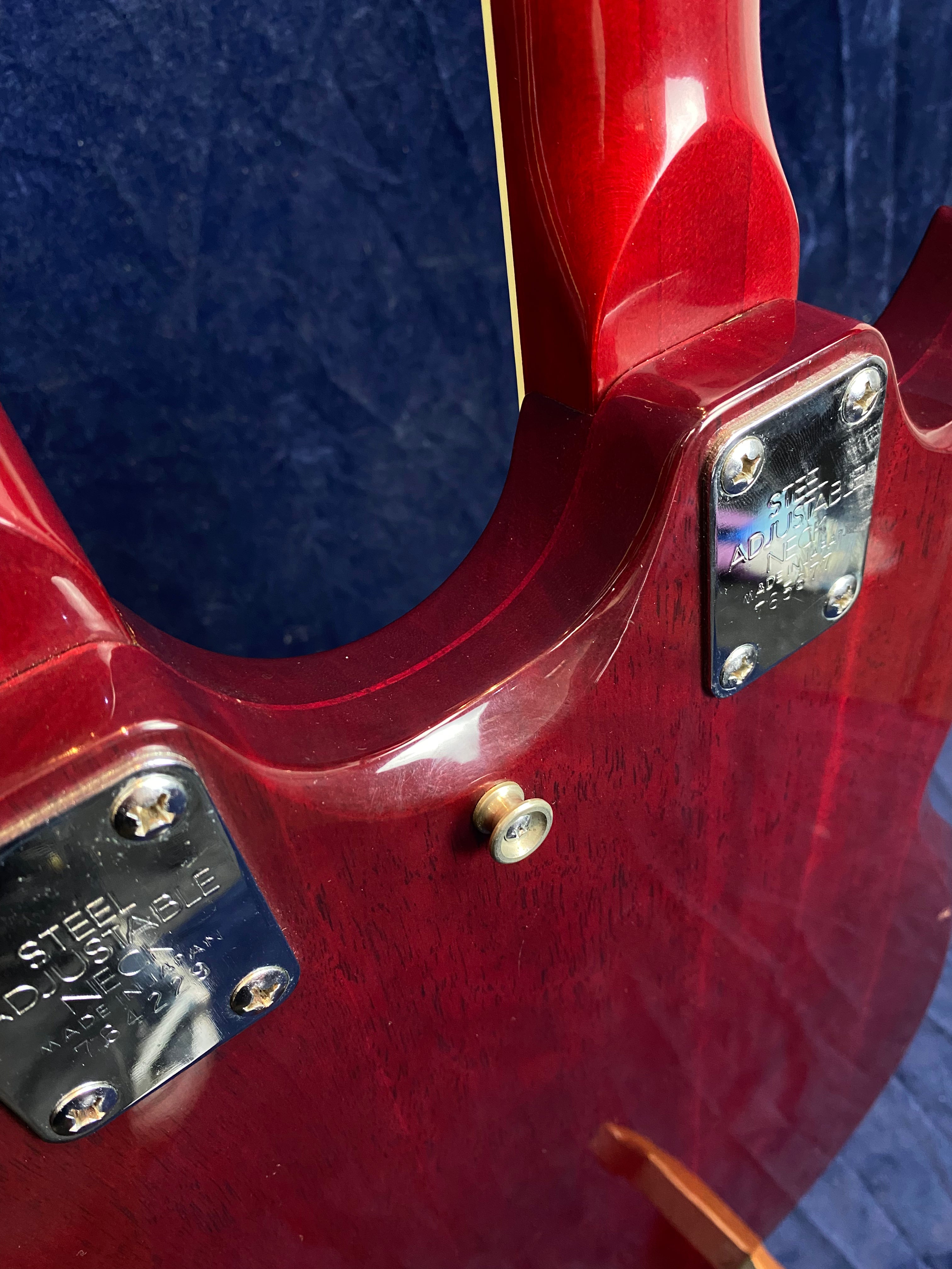 Close-up of a red electric guitar’s neck joint, showing shiny metal plates labeled “Steel Adjustable Neck” and a strap button on the wooden body.