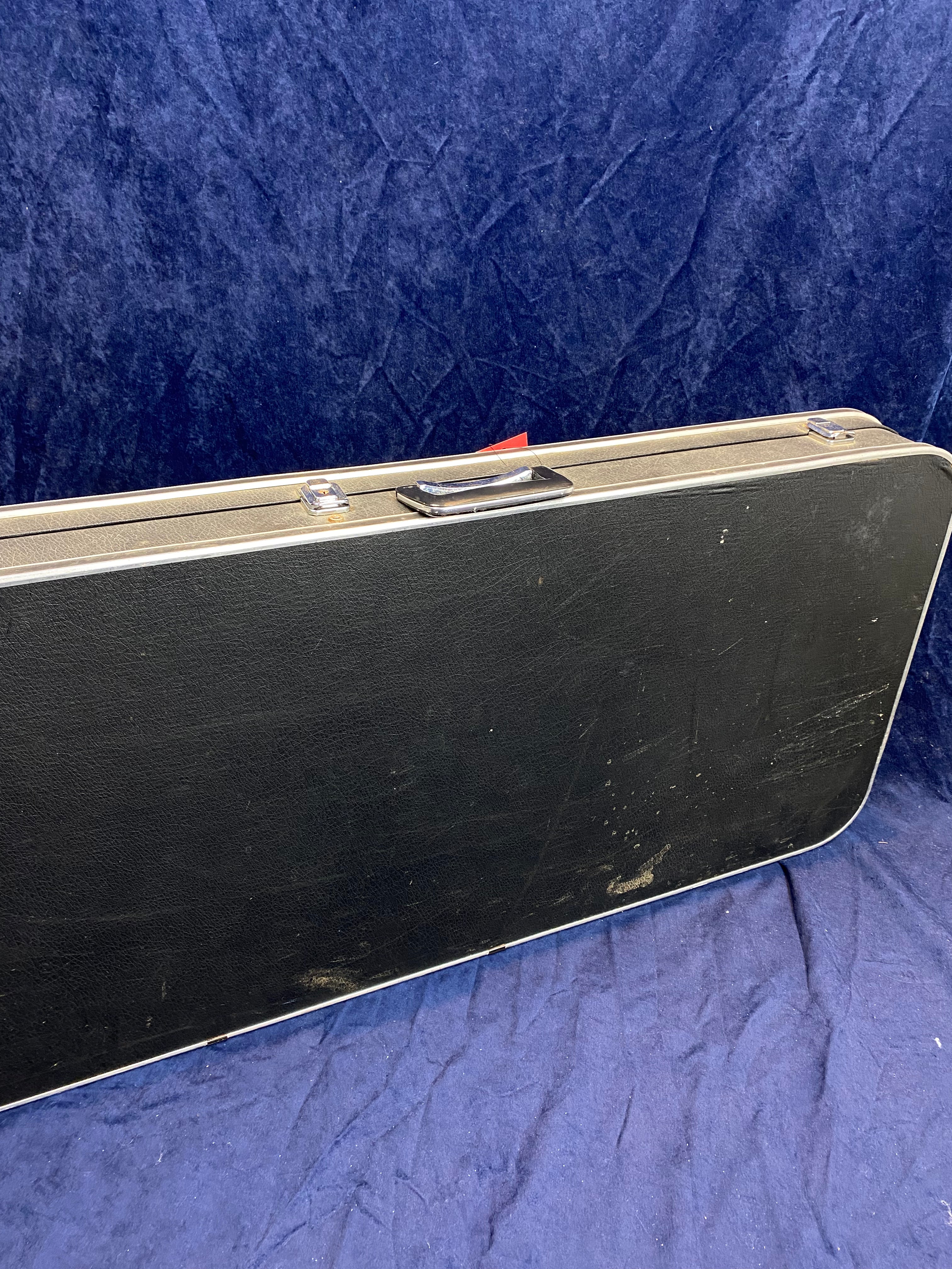 Rectangular black hard guitar case with metal edges and handle, showing some scuffs and scratches, resting on a dark blue textured cloth.