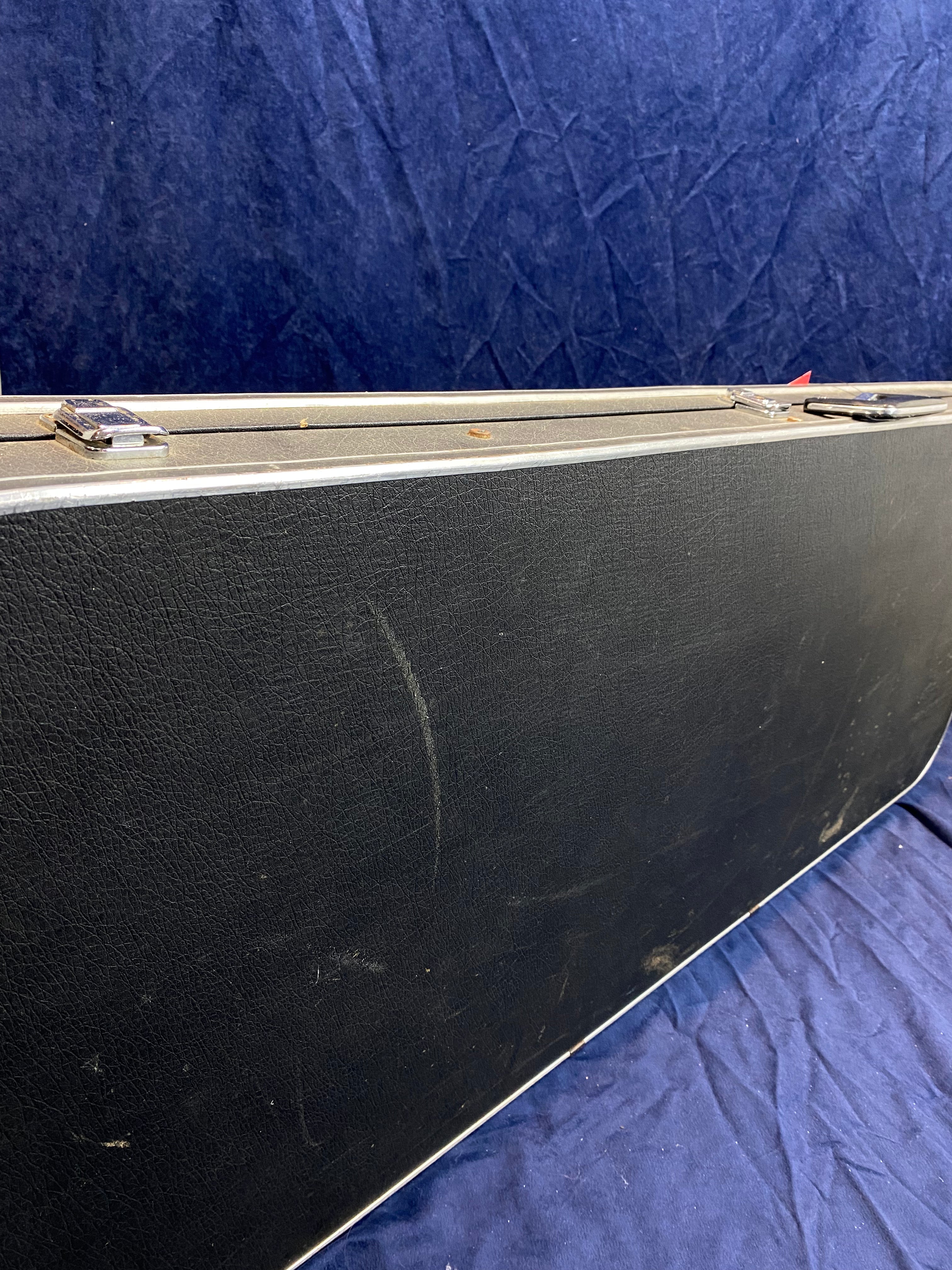 Rectangular black hard guitar case with metal latches, showing scuffs and scratches on a dark blue textured fabric background.