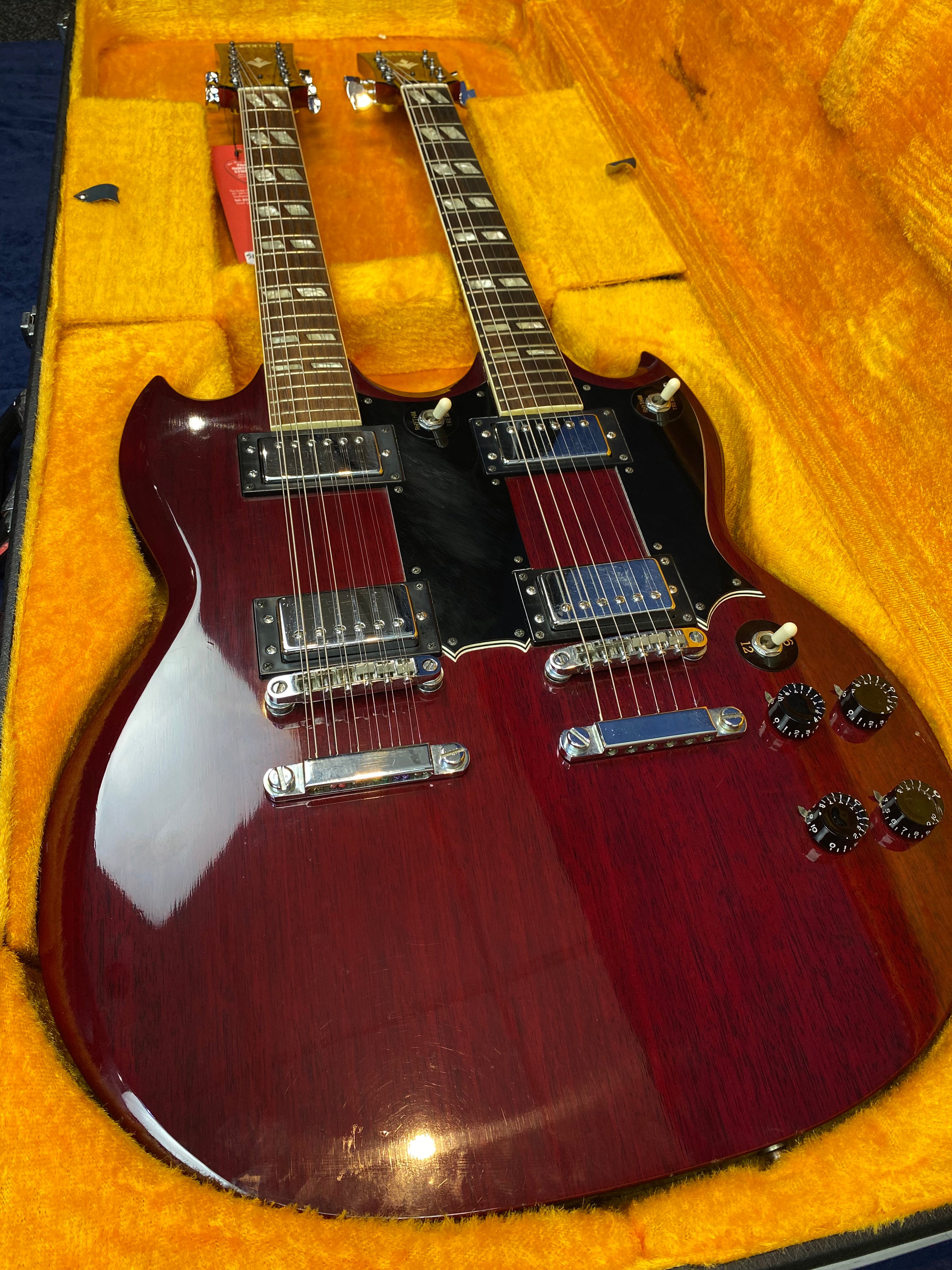Double-neck electric guitar with a glossy cherry red finish, silver hardware, black pickguards, and four control knobs, resting in a yellow plush-lined case.