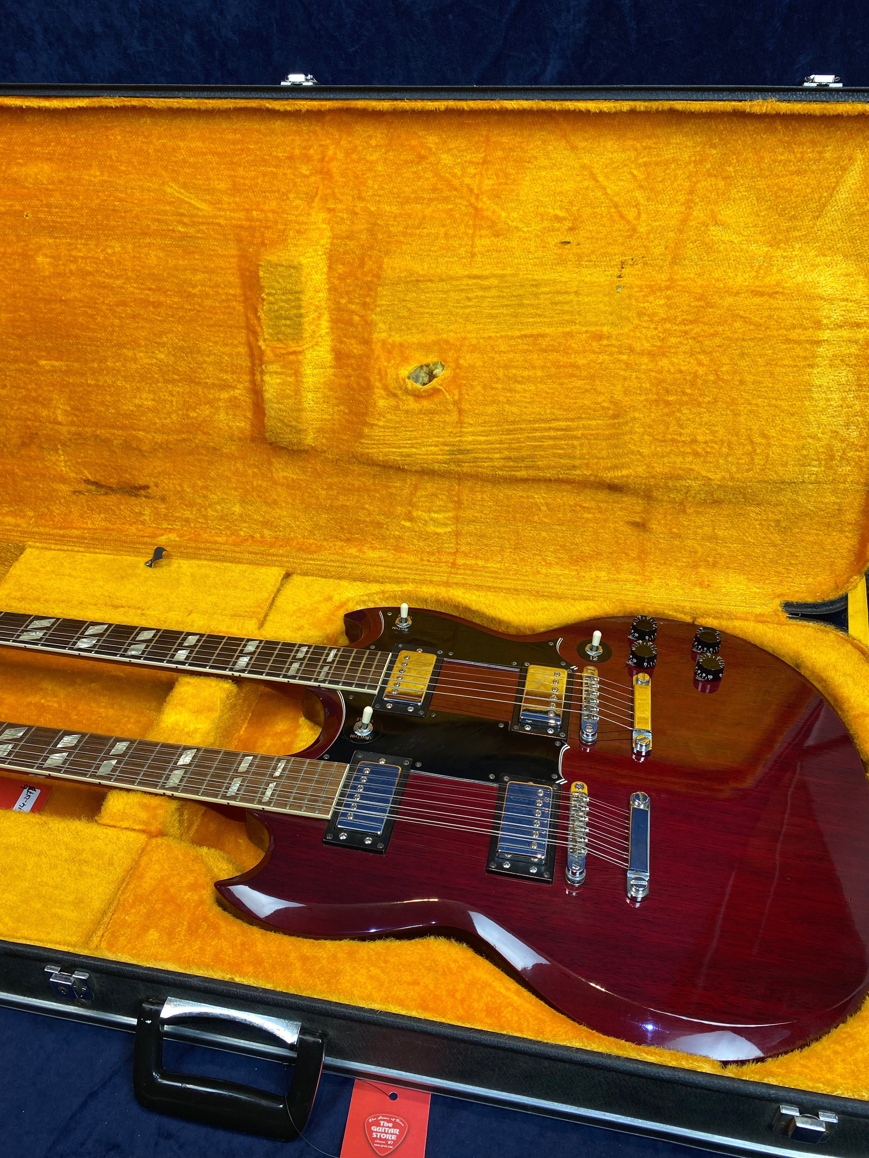 Double-neck cherry red electric guitar with two sets of pickups and controls, resting inside a yellow plush-lined hard case.
