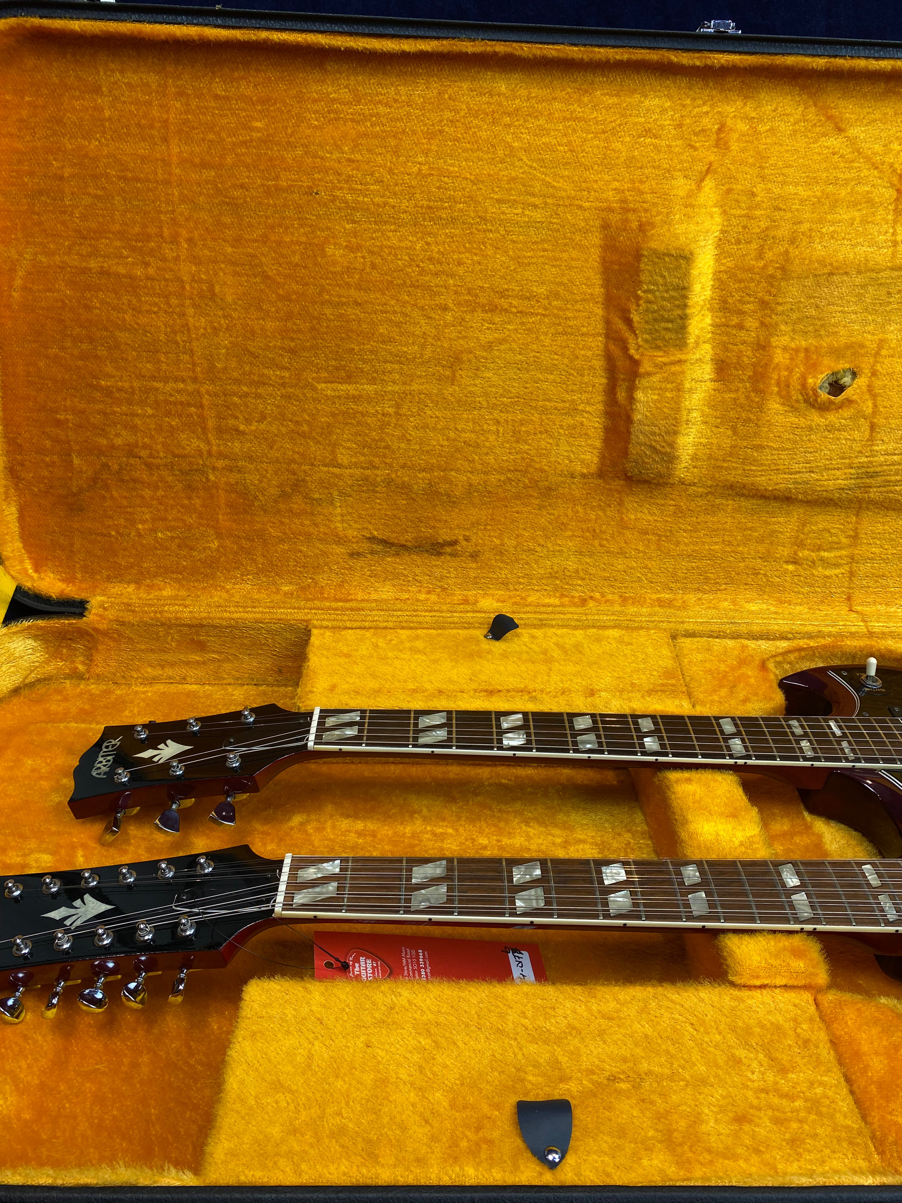 A double-neck electric guitar with two sets of tuning pegs rests in a yellow plush-lined hard case, with two black guitar picks lying nearby.