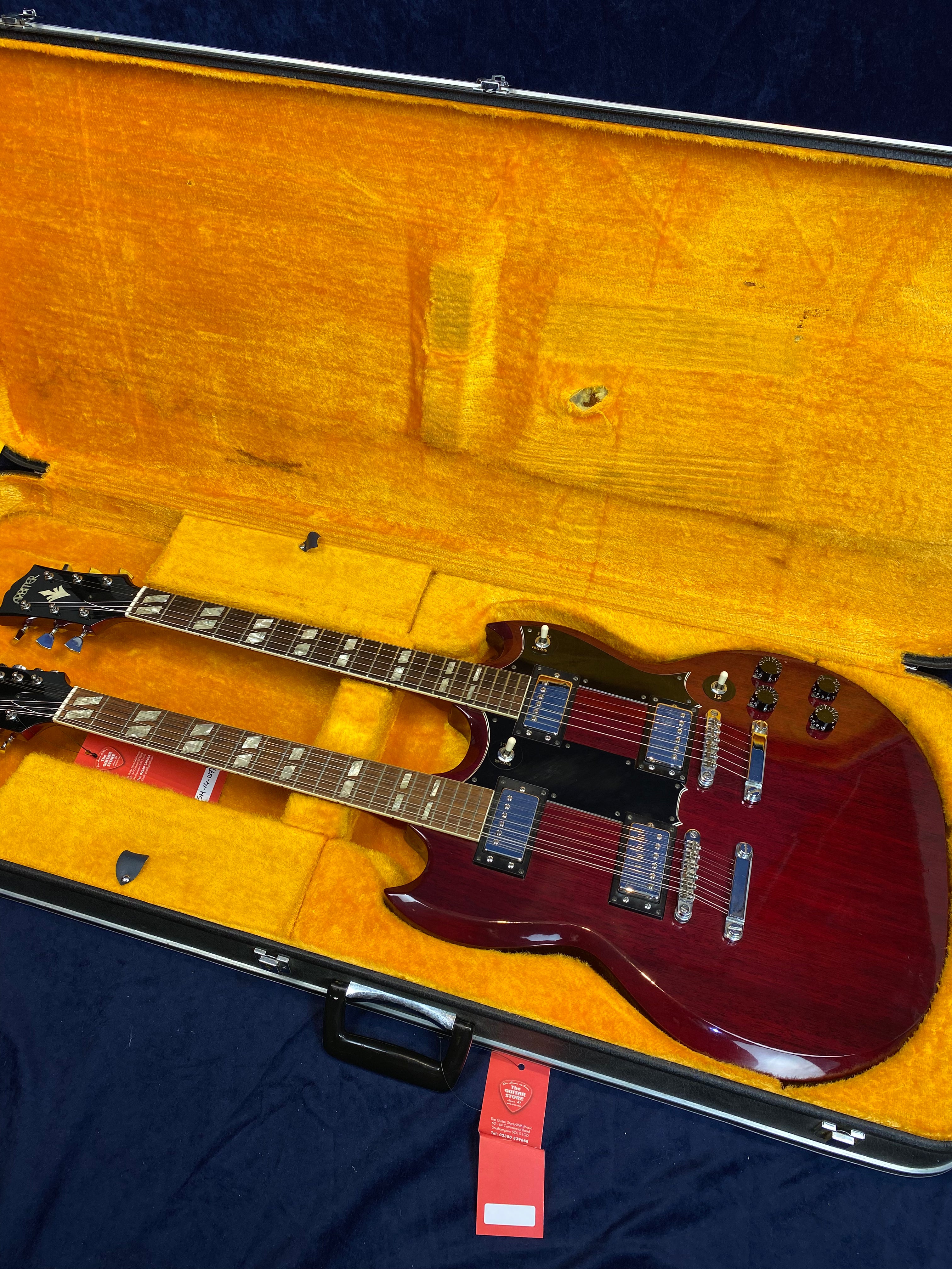 A cherry red double-neck electric guitar with chrome hardware sits in a plush yellow hard case, with a red tag attached to the handle.