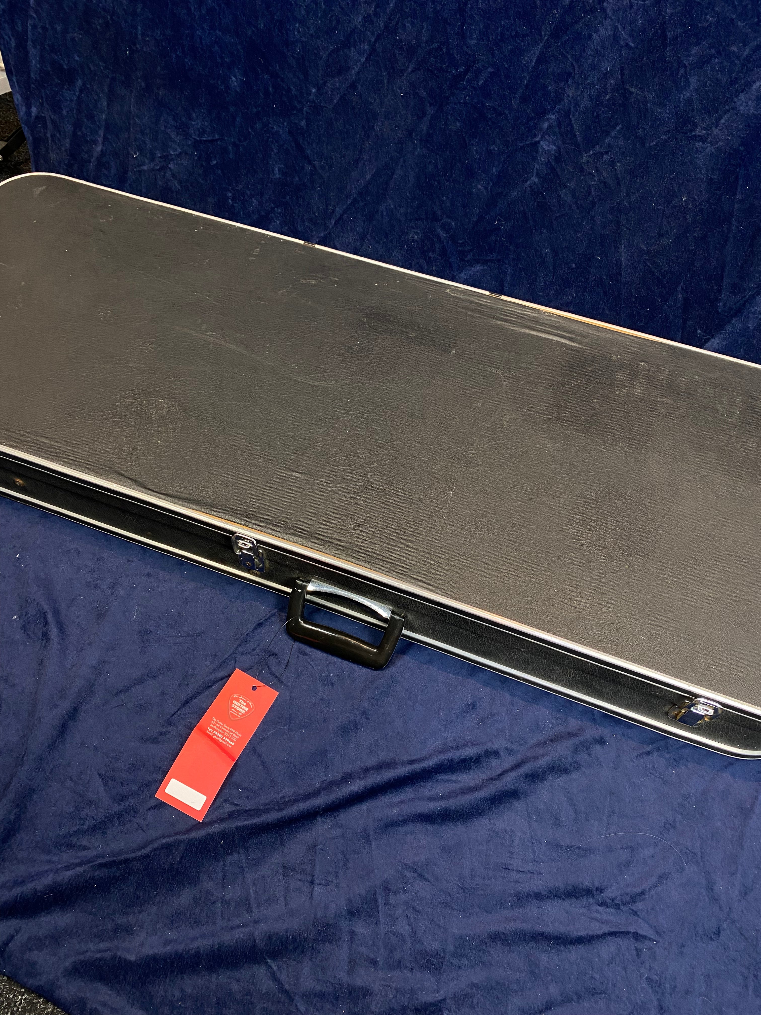 Rectangular black hard case with a handle for an electric guitar, resting on a blue cloth, red tag attached near the handle.