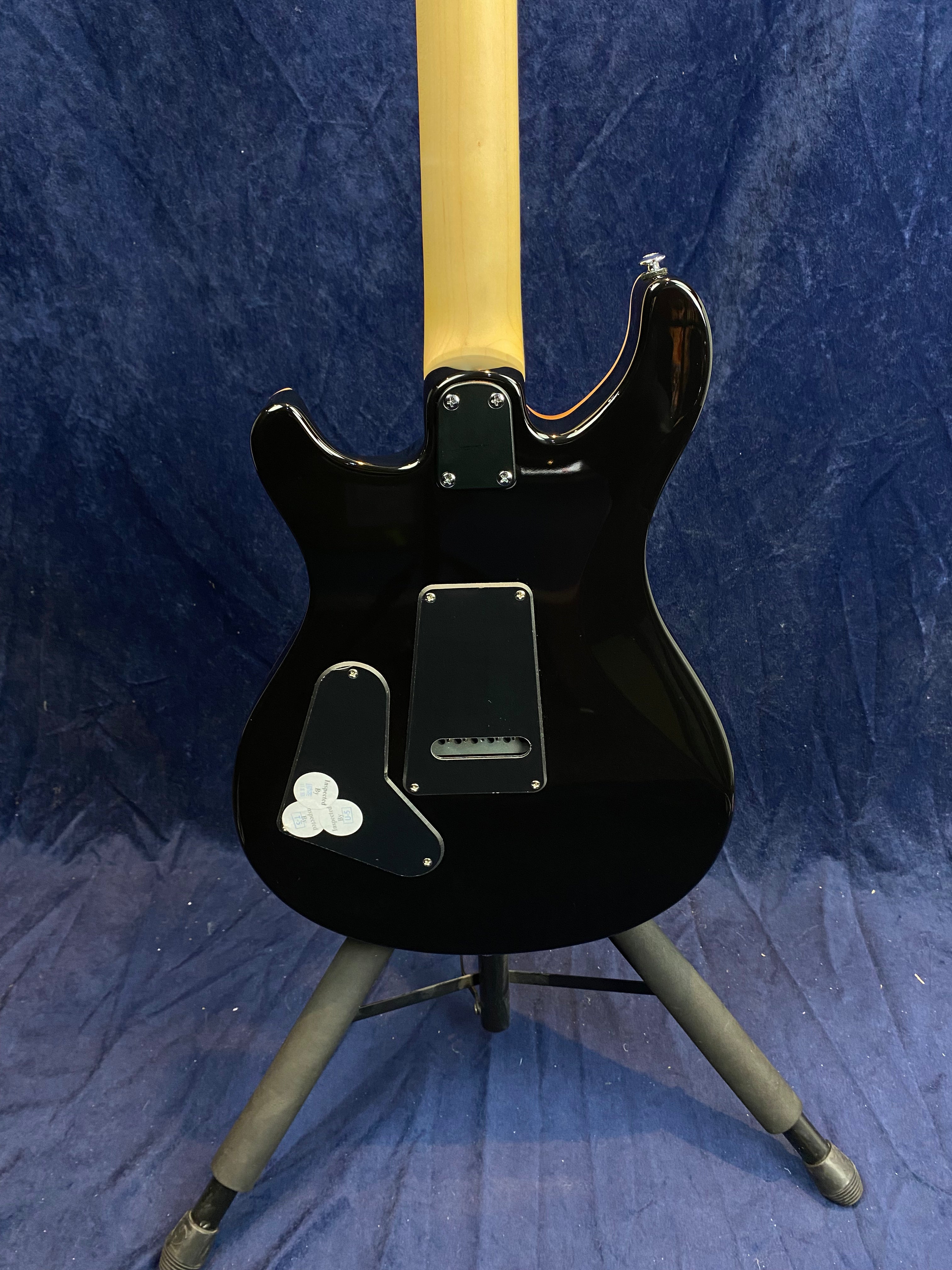 Black electric guitar shown from the back, with a natural wood neck, resting on a stand, and three guitar picks stuck to a cover plate.