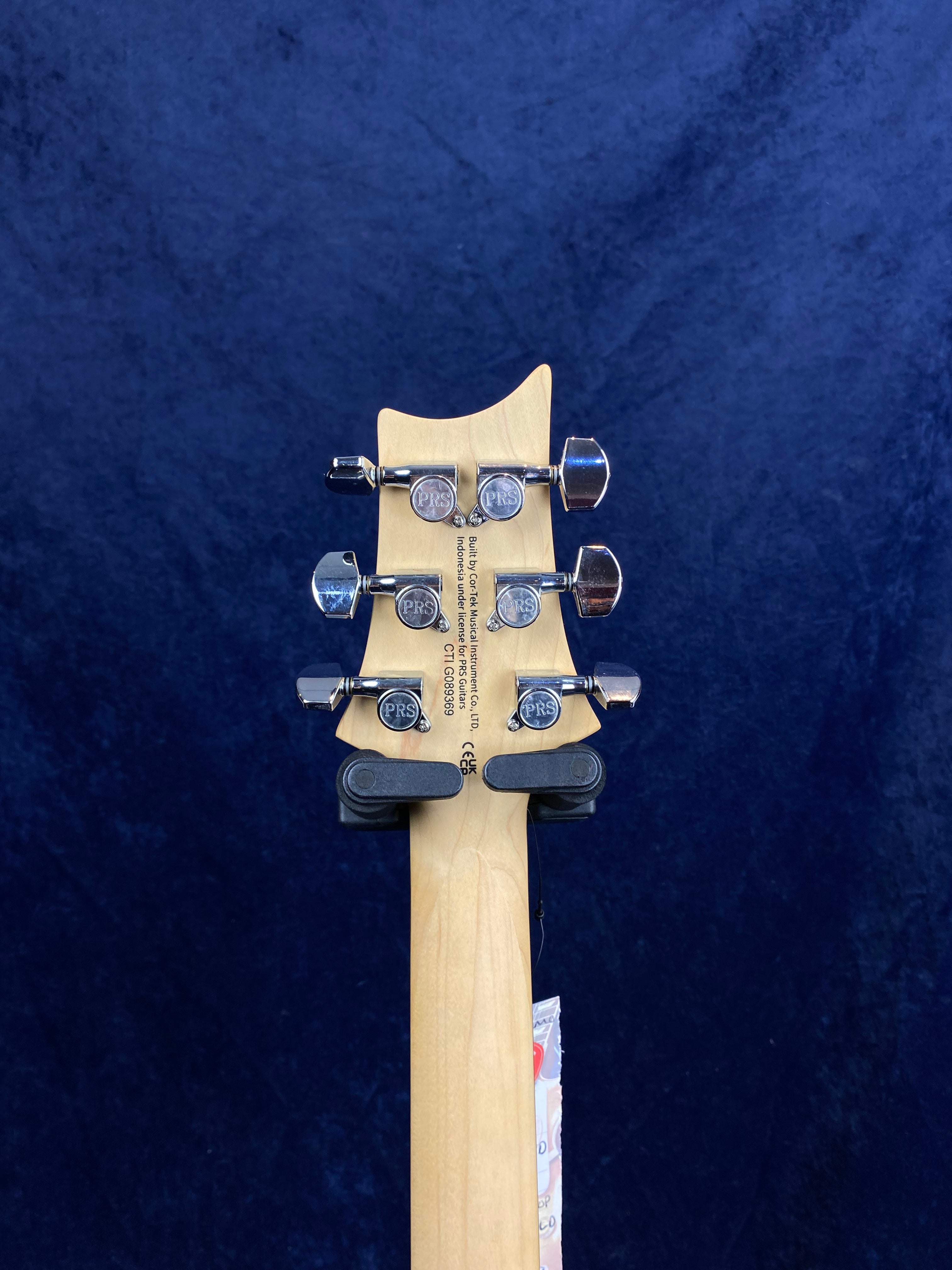 Light wood electric guitar headstock with six chrome tuners, each marked "PRS," against a dark blue background.