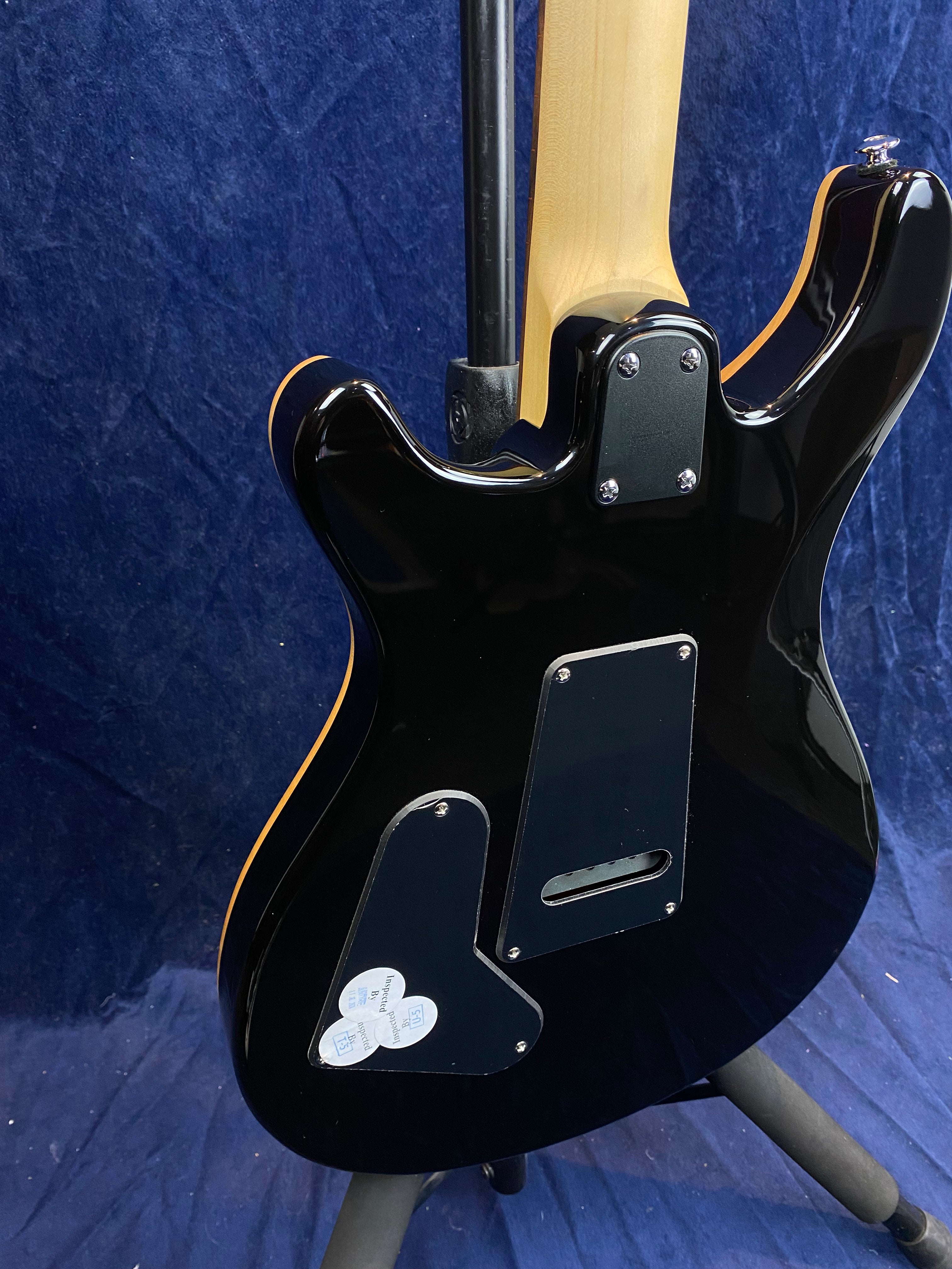 Black electric guitar back view, revealing multiple control cavity covers and a light wood neck joint with visible screws.