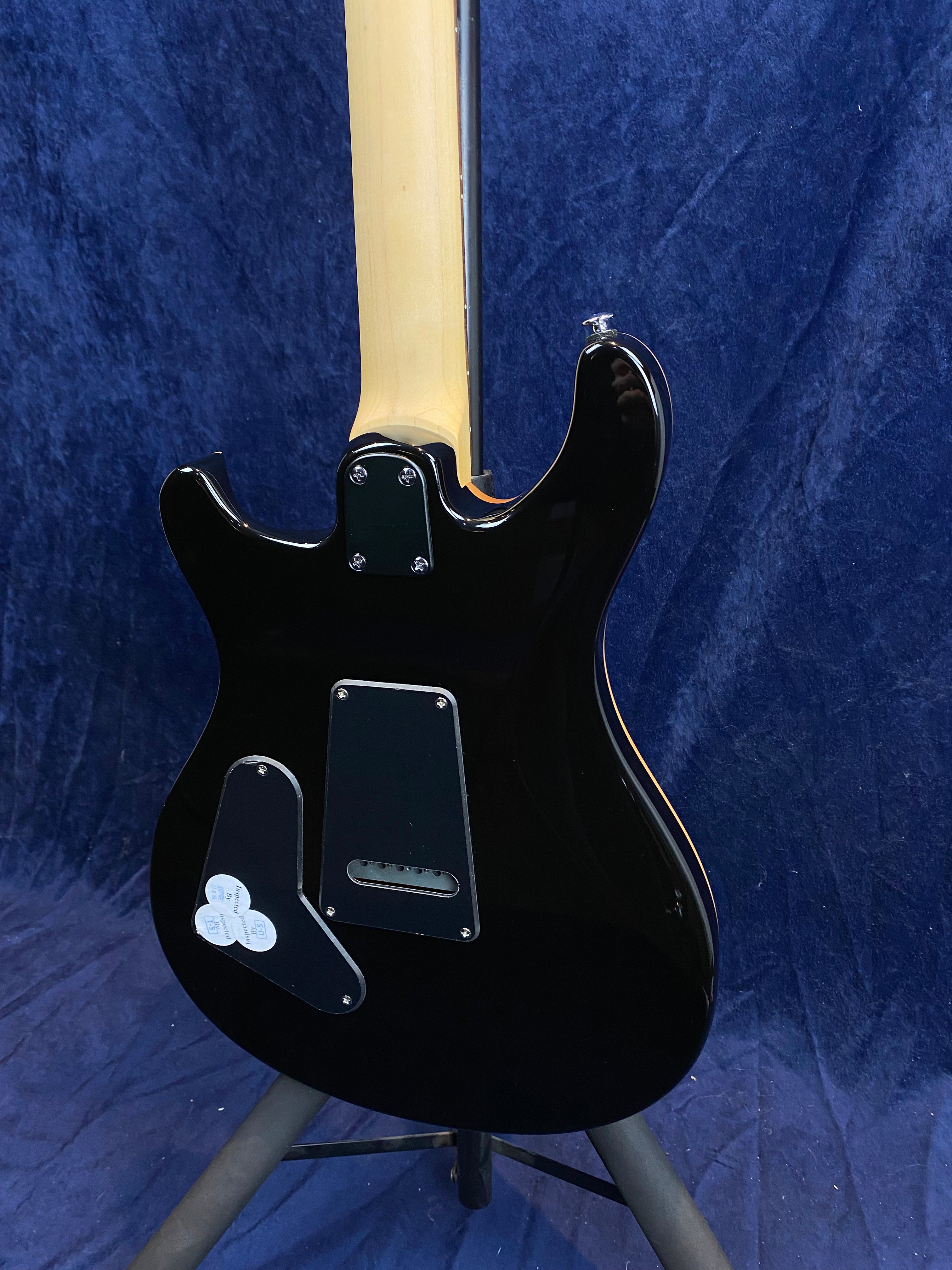 Black electric guitar viewed from the back, showing the light wood neck and back cavity plates against a dark blue background.