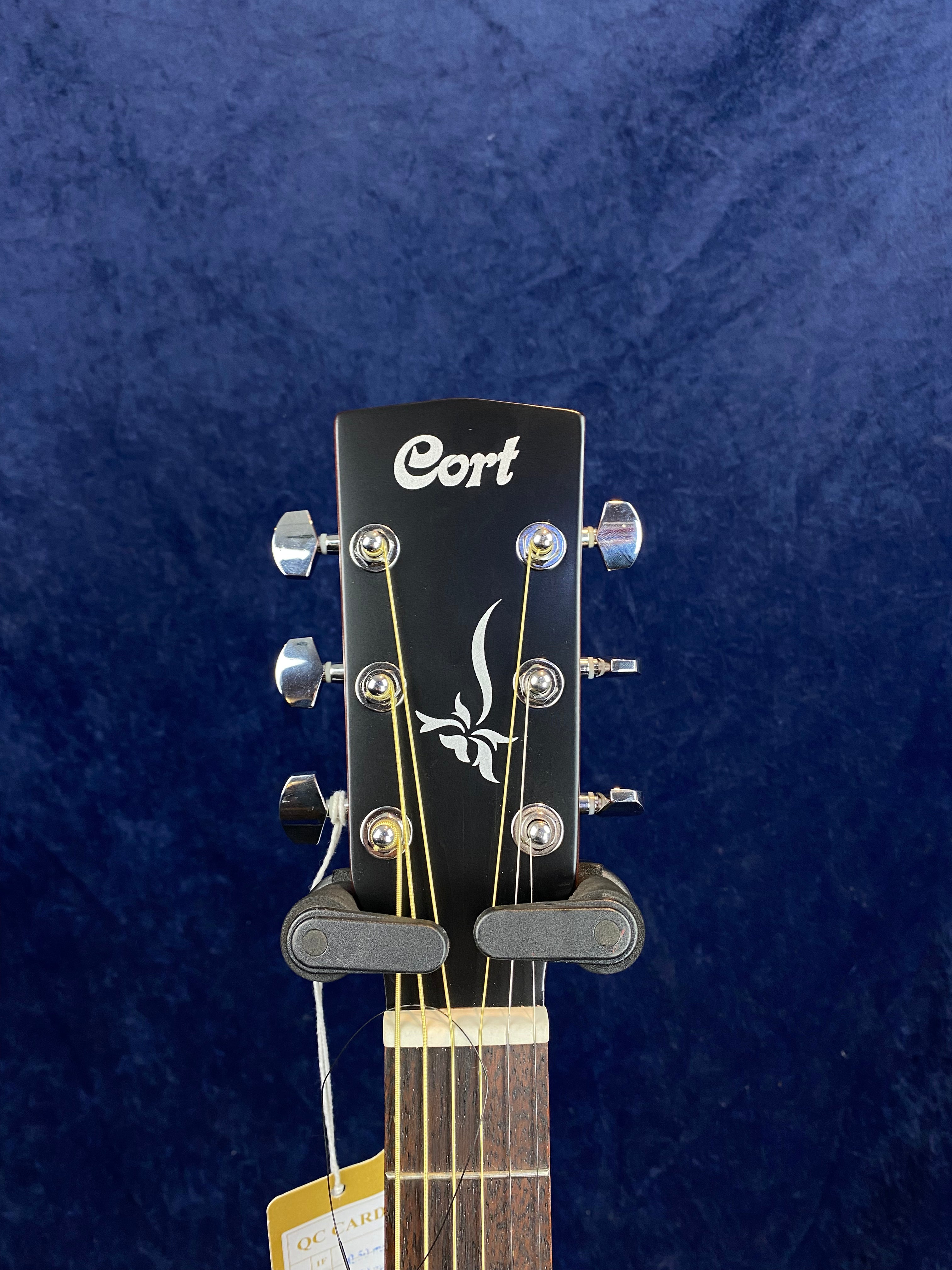 Tête de guitare acoustique Cort noire avec six mécaniques chromées, décor végétal blanc au centre, étiquette suspendue au manche.