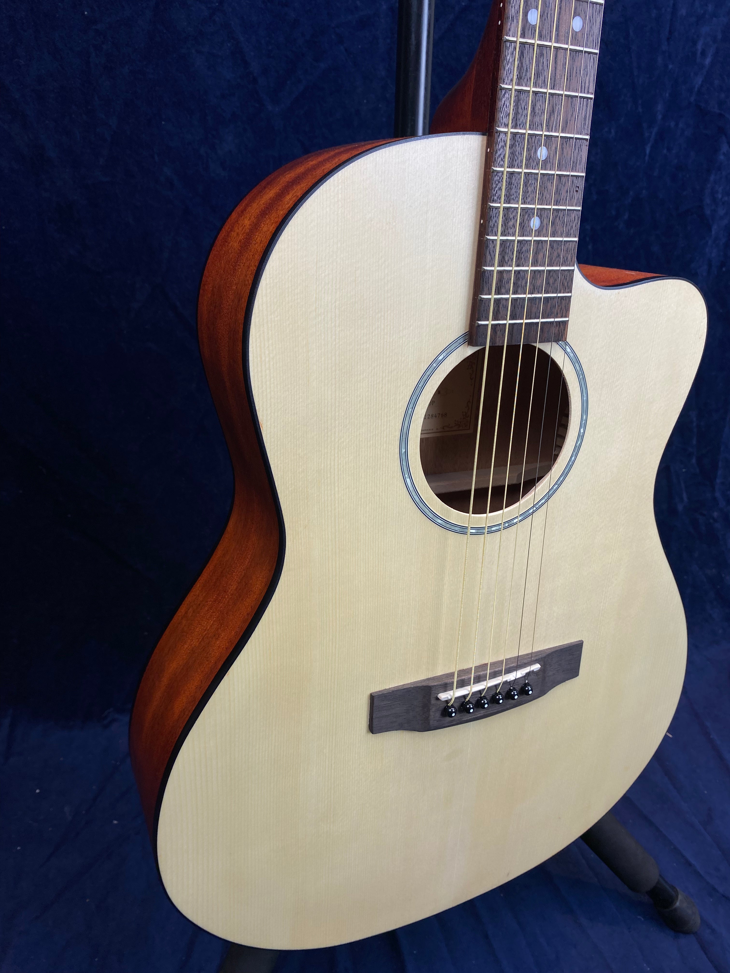 Light wood acoustic guitar with a cutaway body, dark brown sides, round soundhole, and six steel strings, displayed upright against a dark background.