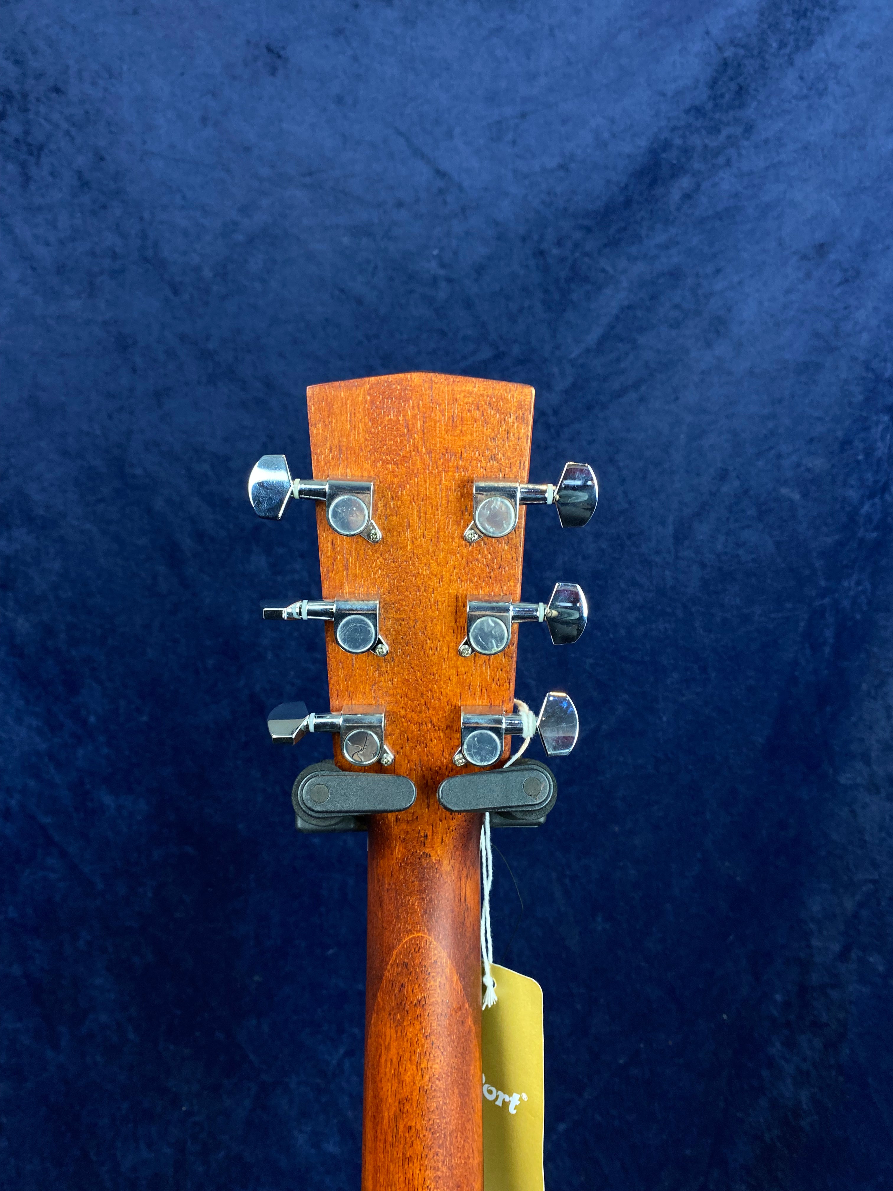 Back view of an acoustic guitar headstock showing six metal tuning pegs, wooden neck, and a brown tag hanging from the tuning hardware.