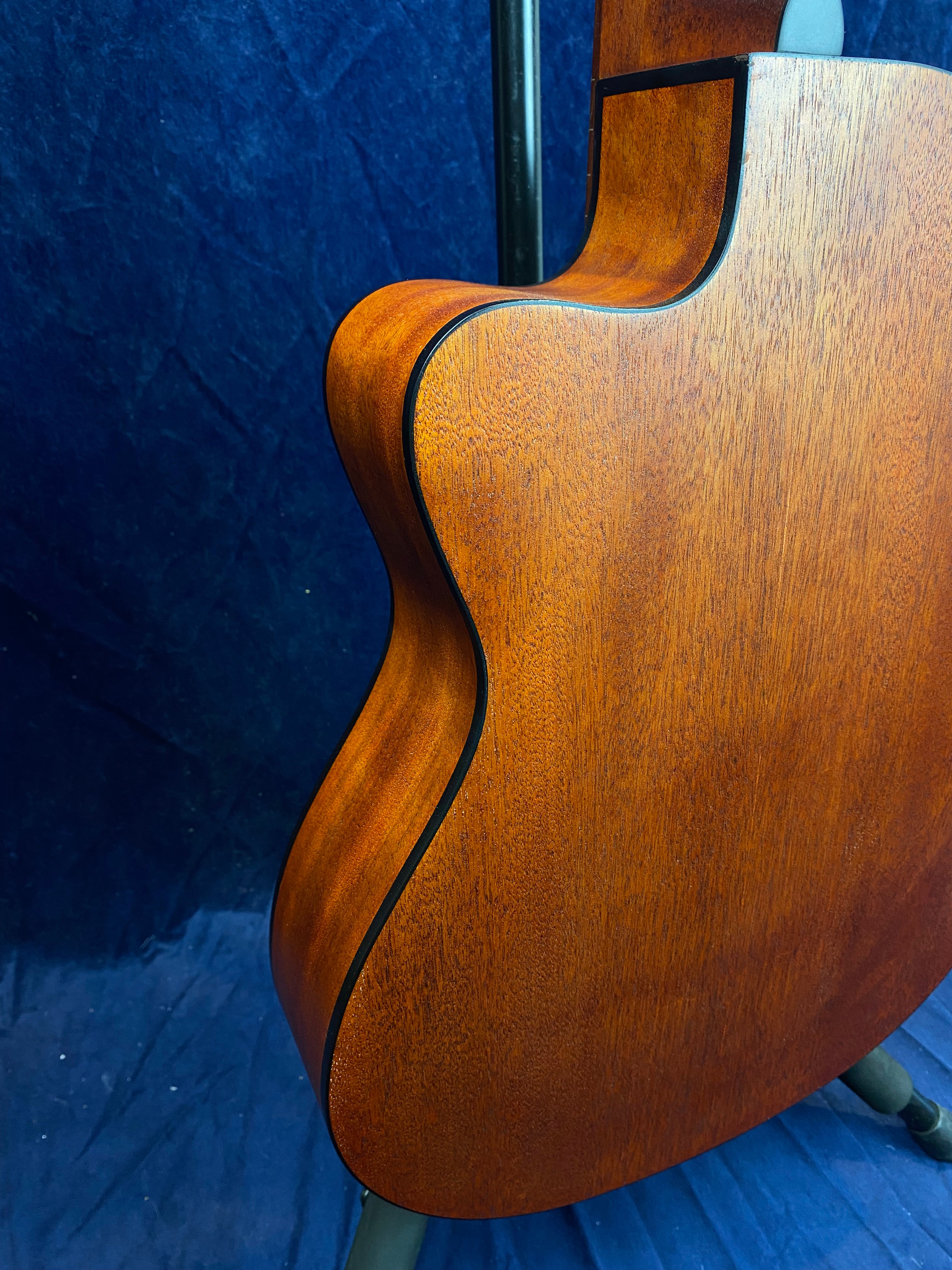 Warm brown acoustic guitar body with a cutaway design, smooth matt wood grain, and black edging, shown from the back against a dark blue background.