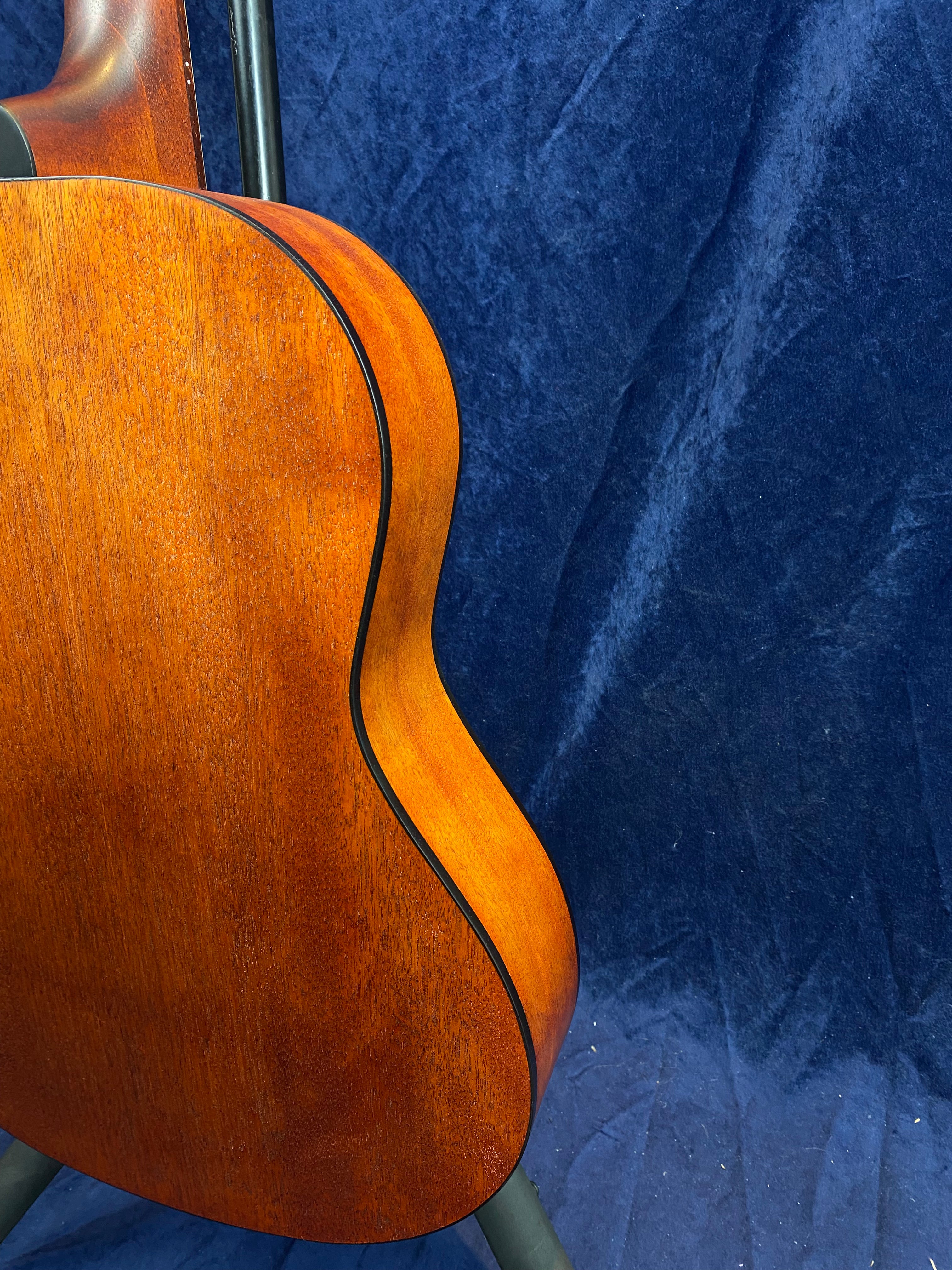 Curved wooden body of an acoustic guitar with a smooth, warm brown finish and black trim, shown against a dark blue fabric background.