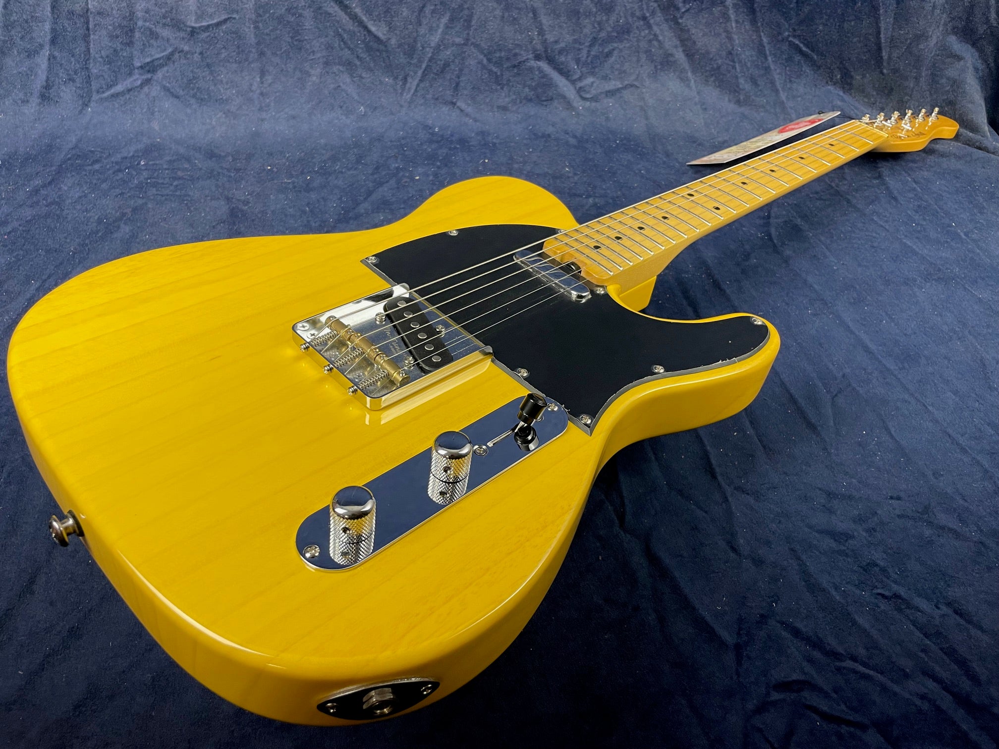 Yellow electric guitar with a black pickguard, chrome knob controls, single cutaway body, and maple neck laying on a dark fabric background.