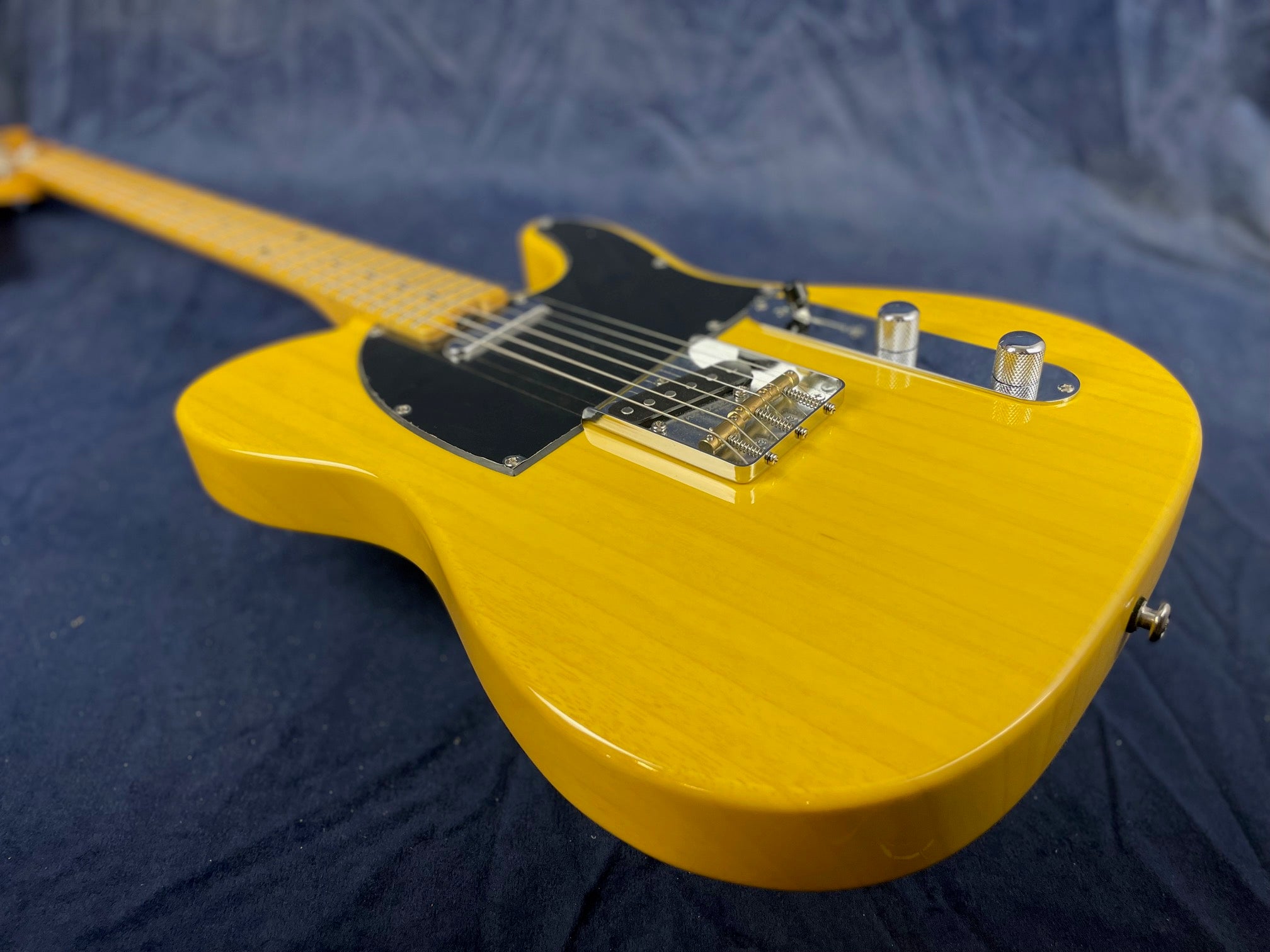 Yellow electric guitar with a black pickguard, chrome bridge, and two metal control knobs on a dark fabric background.