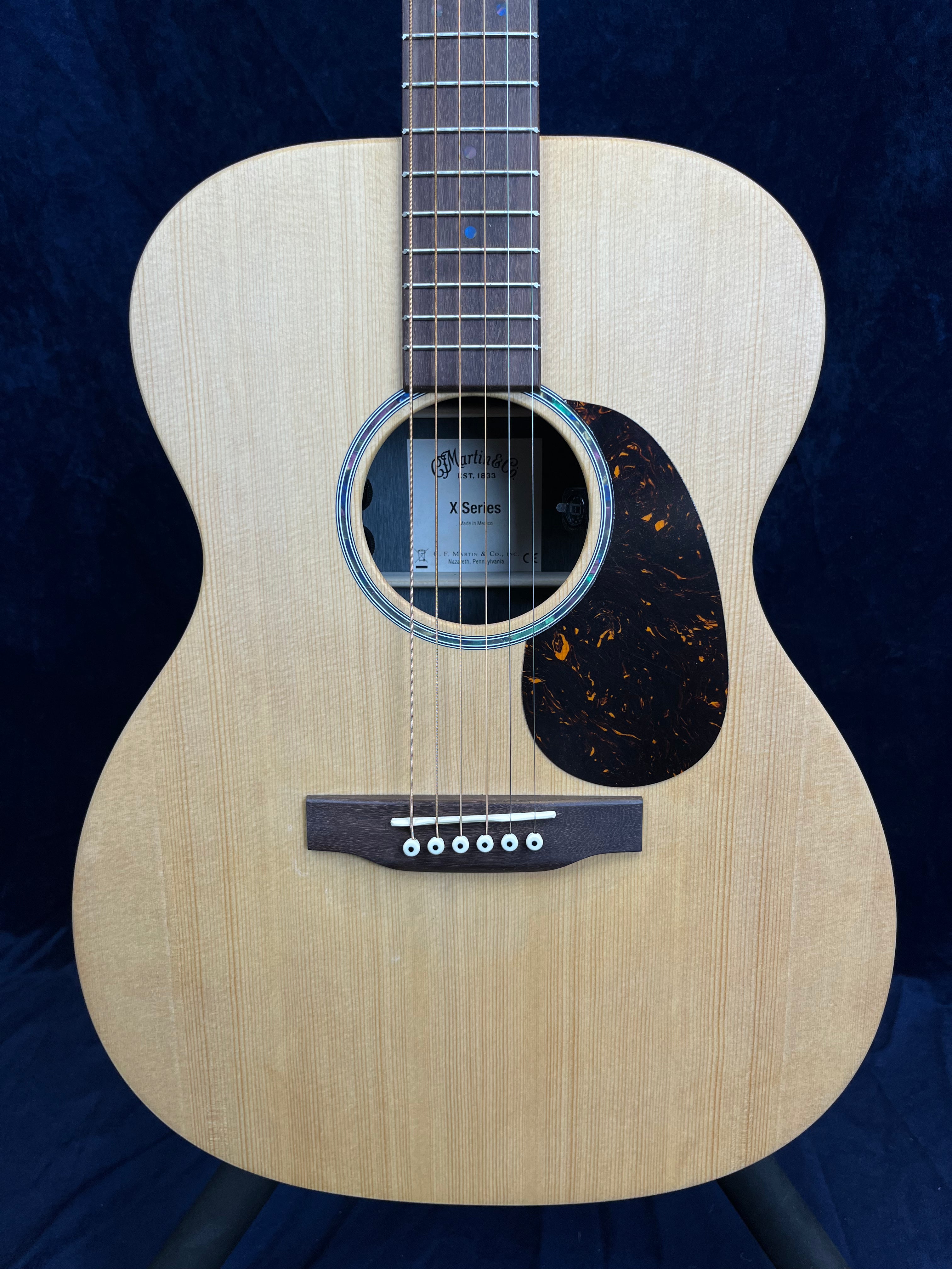 Light wood acoustic guitar with a round sound hole, dark pickguard, and six white-peg bridge pins; label inside reads "Martin & Co X Series."