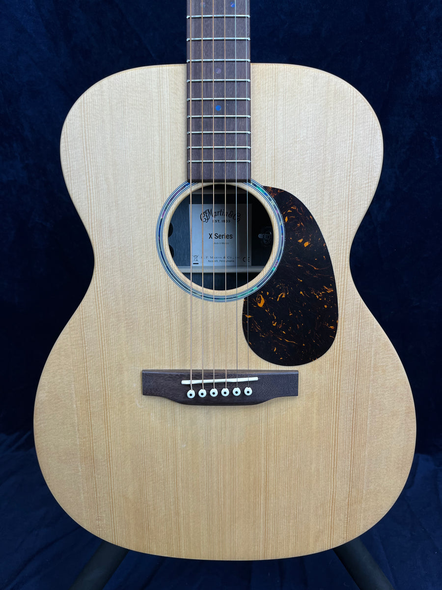Light wood acoustic guitar with a round sound hole, dark pickguard, and six white-peg bridge pins; label inside reads "Martin & Co X Series."