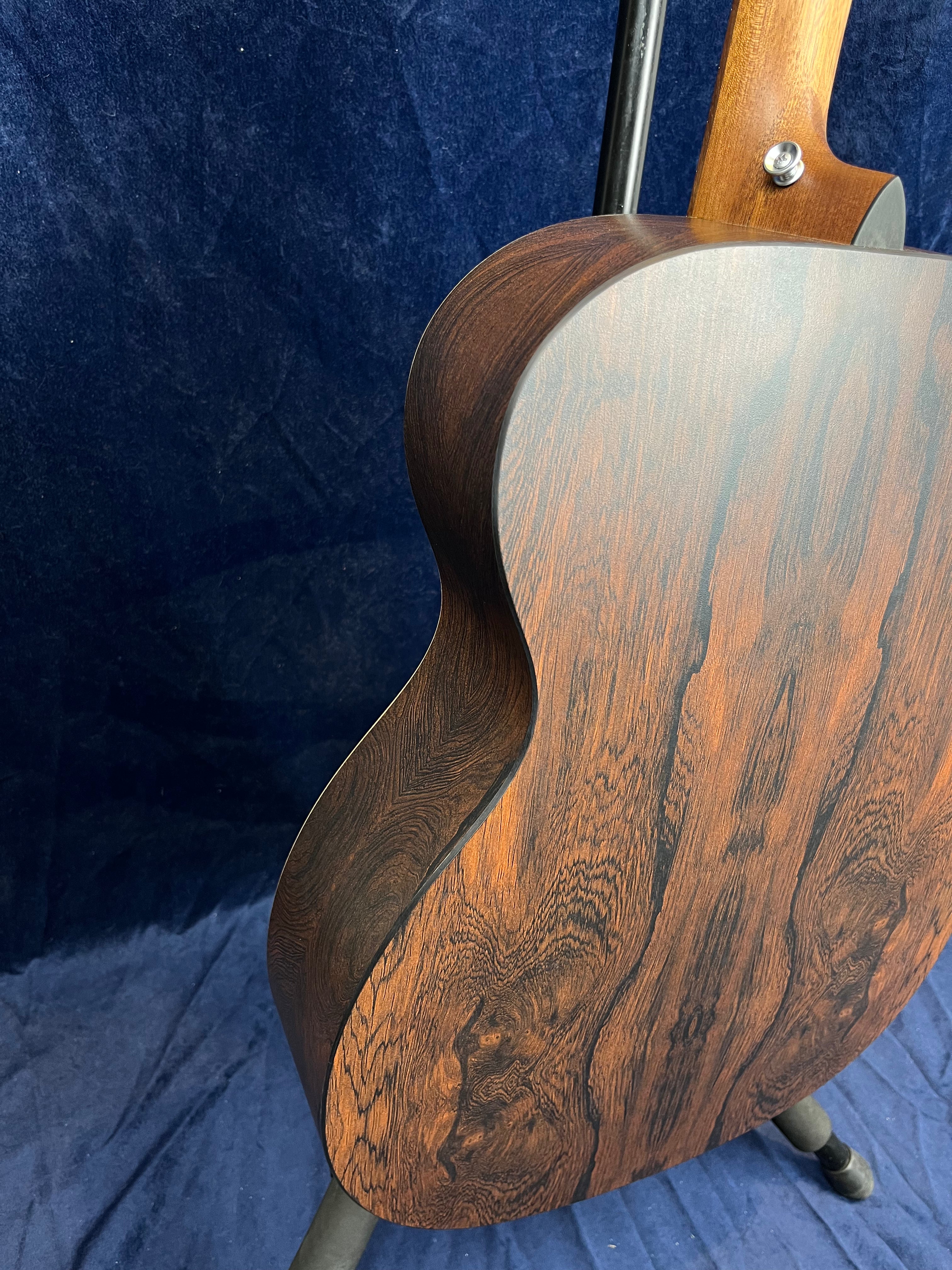 The back of an acoustic guitar with dark brown wood grain patterns, shown close up while resting on a stand against a dark blue fabric background.