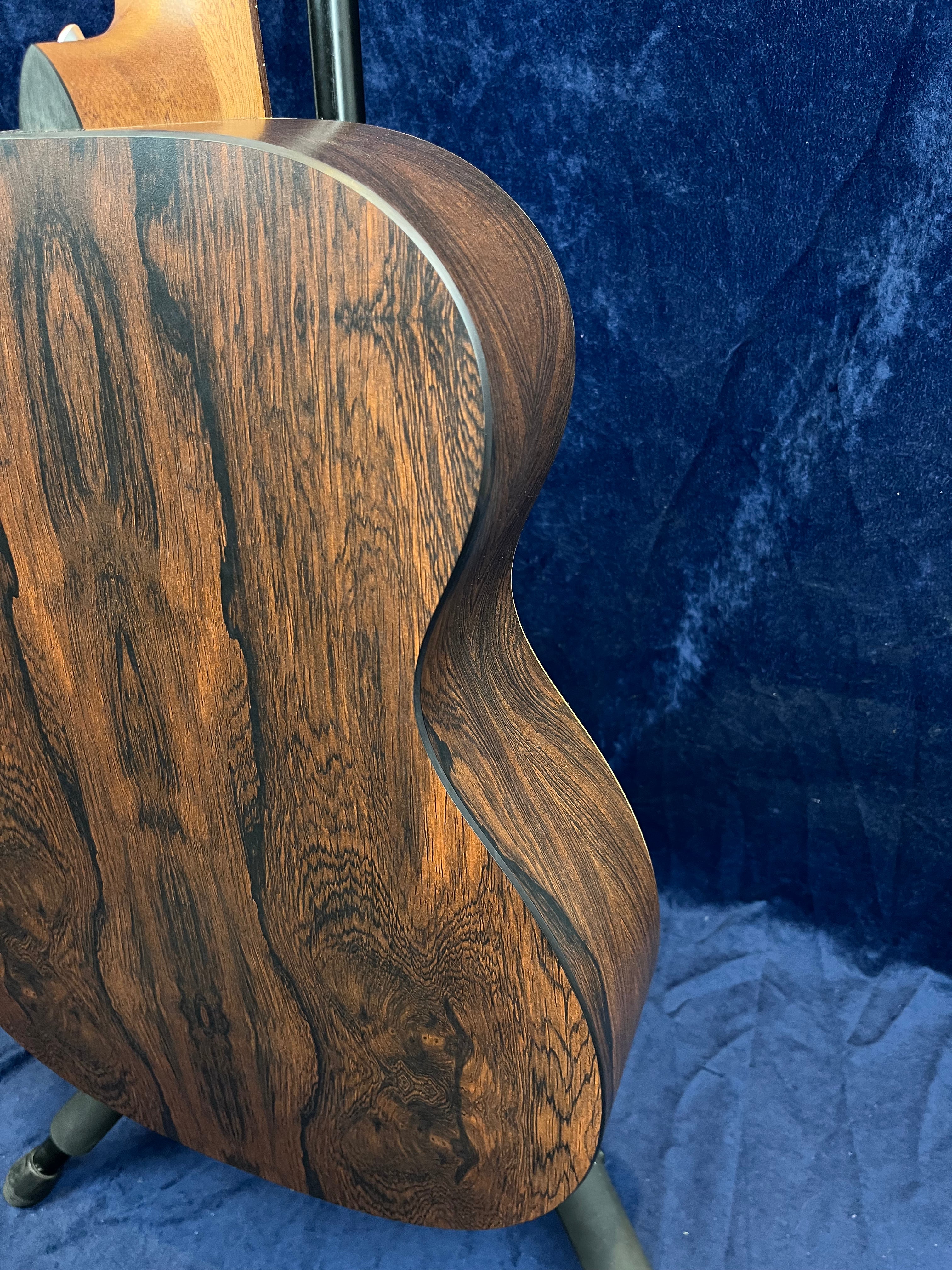 Curved back and side of an acoustic guitar in dark brown wood with striking black grain patterns, set against a dark blue textured background.