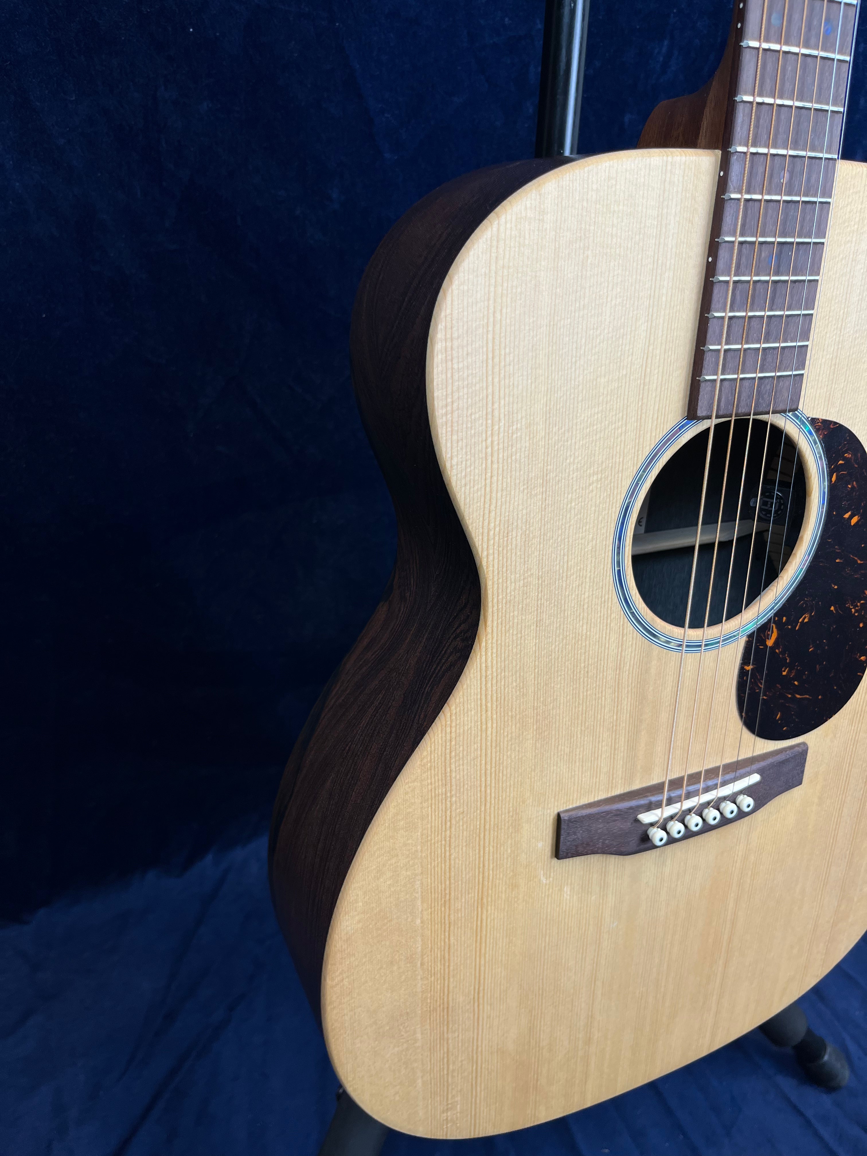 Acoustic guitar with a light wood top, dark wood sides, tortoiseshell pickguard, and six strings, shown on a stand against a dark background.