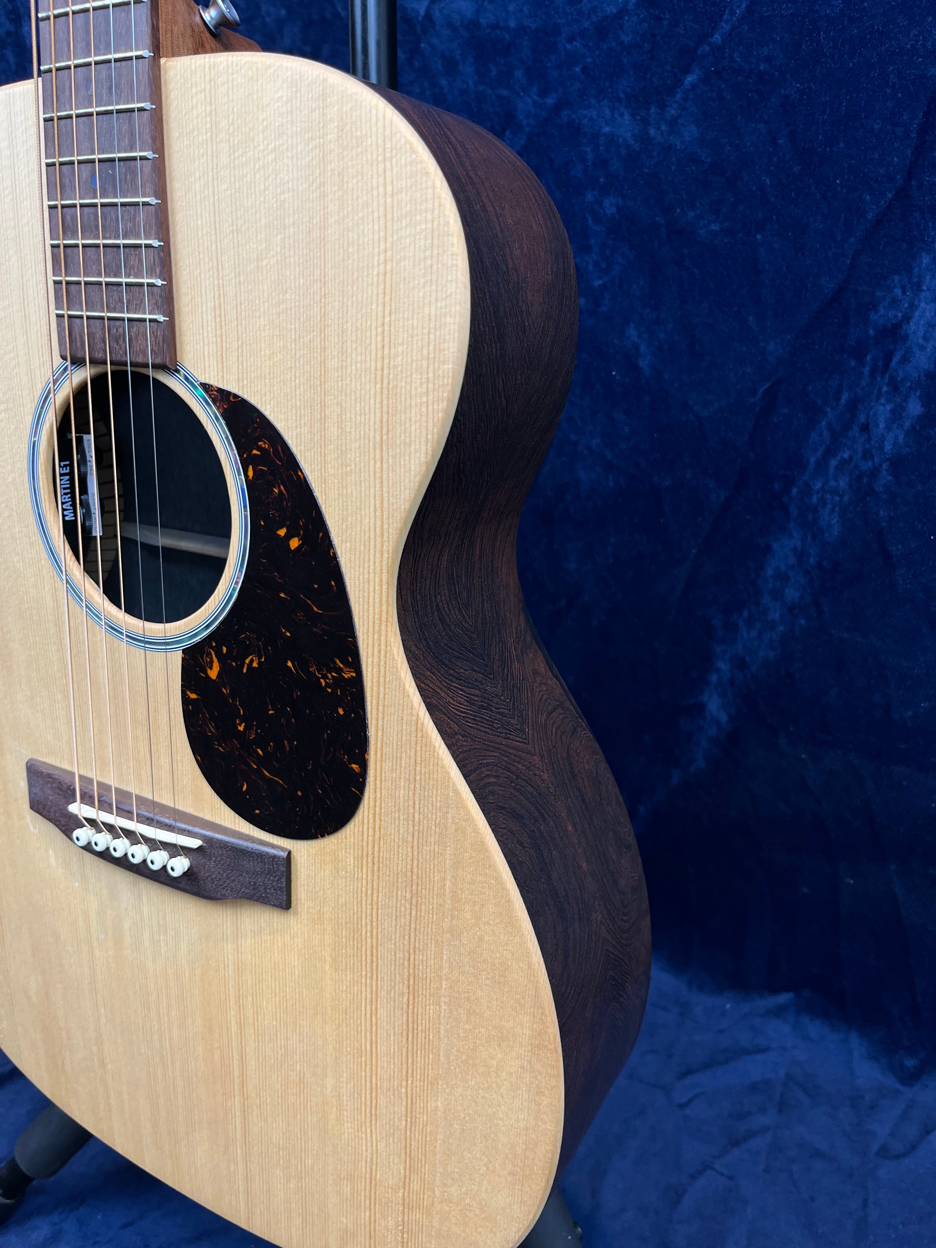 Acoustic guitar with a light wood top, dark wood sides, tortoise-patterned pickguard, and six strings against a dark blue textured background.