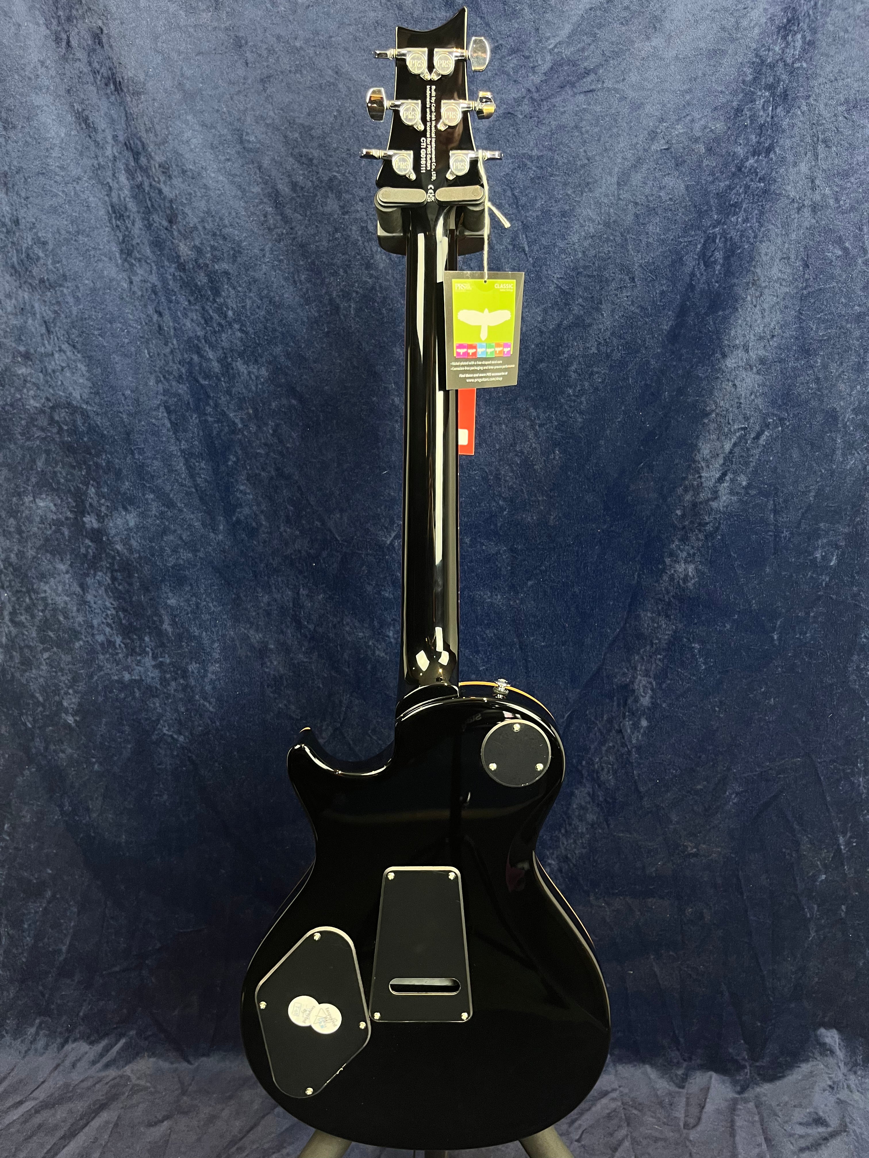 Glossy black electric guitar shown from the back, revealing multiple access panels and chrome tuners against a dark blue textured backdrop.