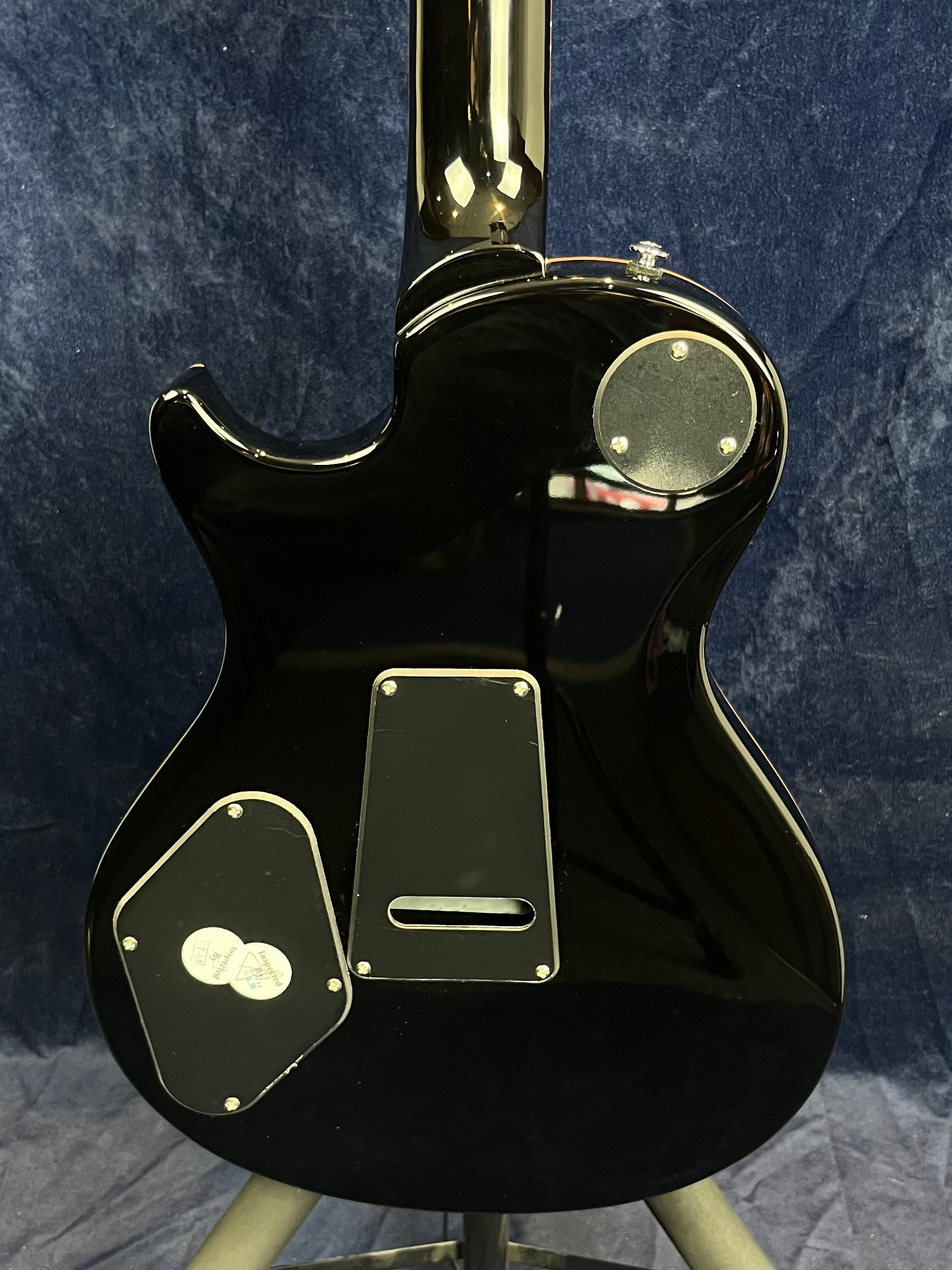 Glossy black electric guitar back, showing three access panels with screws and a rounded single cutaway body shape against a dark textured background.