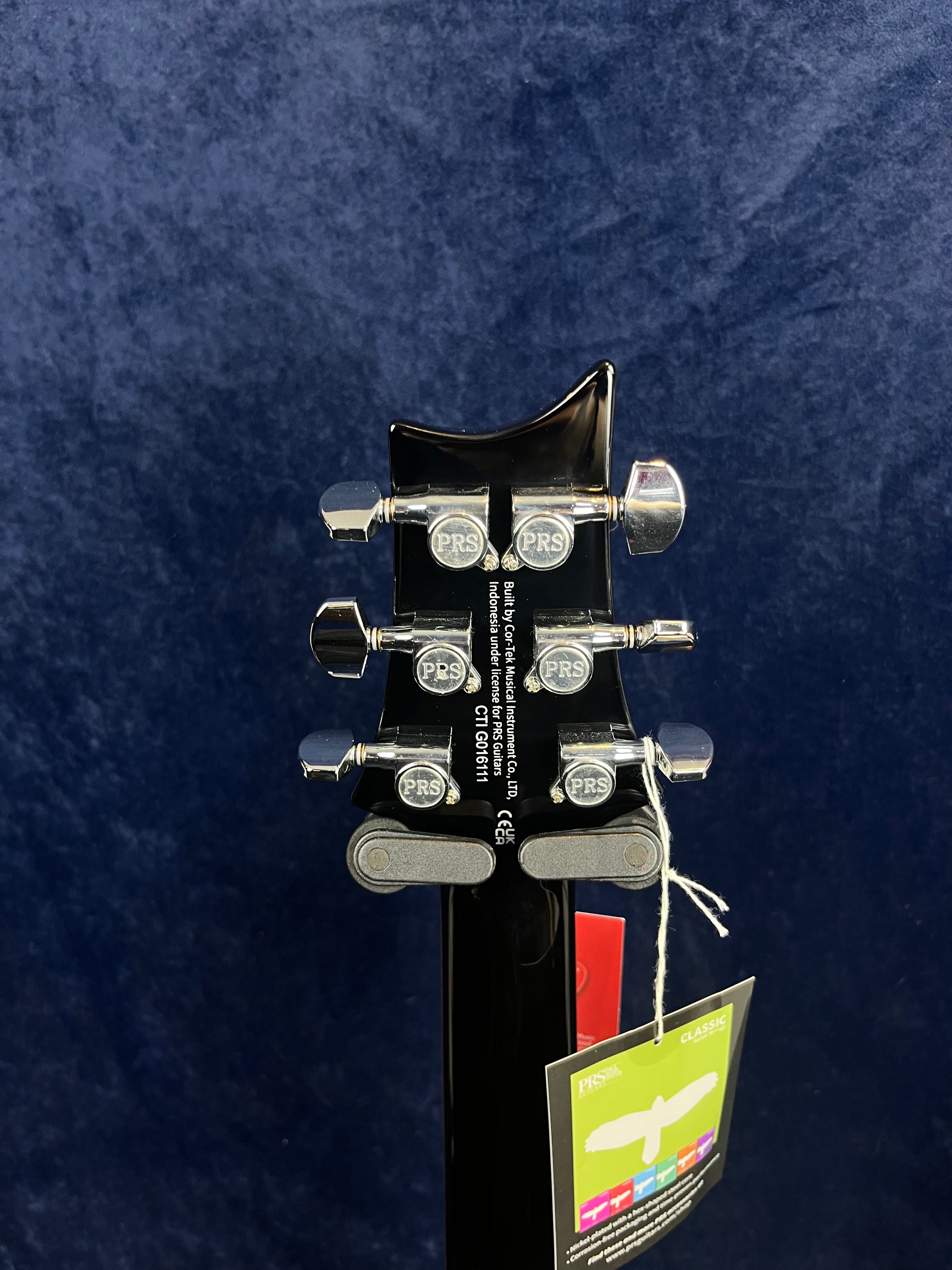 Back of a PRS electric guitar headstock showing six chrome tuning pegs and tags attached to the neck against a dark blue background.