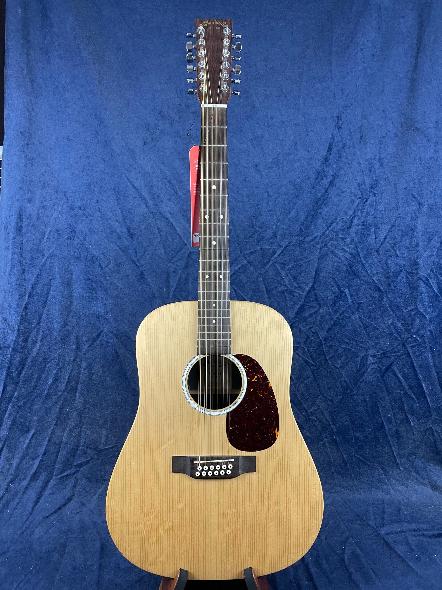 Twelve-string acoustic guitar with a light wood body, dark pickguard, and metal tuning pegs, displayed upright against a blue textured background.