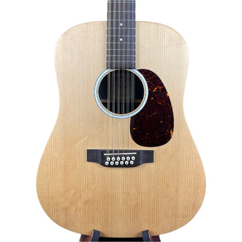Light wood acoustic guitar with a dark brown pickguard, twelve strings, circular sound hole, and white dot inlays on a dark fretboard.